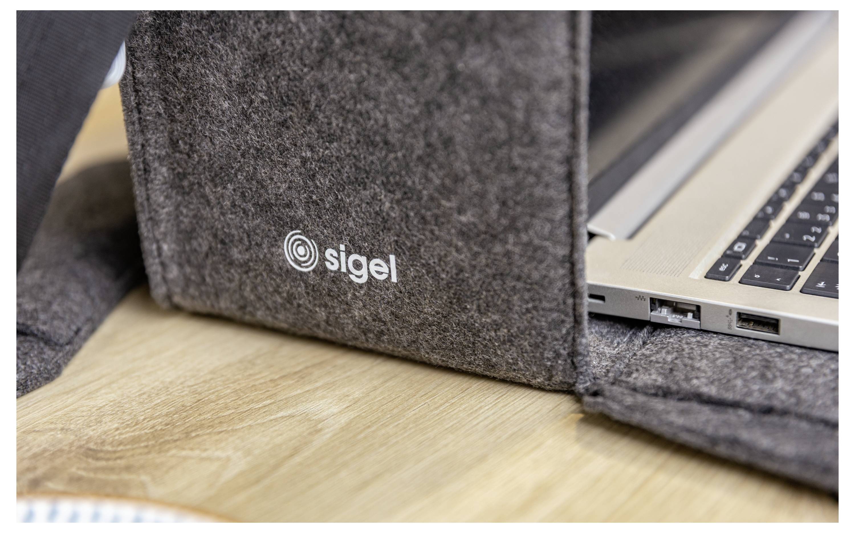 Nahaufnahme einer Filz-Laptop-Schutzhülle mit dem "sigel"-Logo neben einem aufgeklappten Laptop auf einer Holzoberfläche.