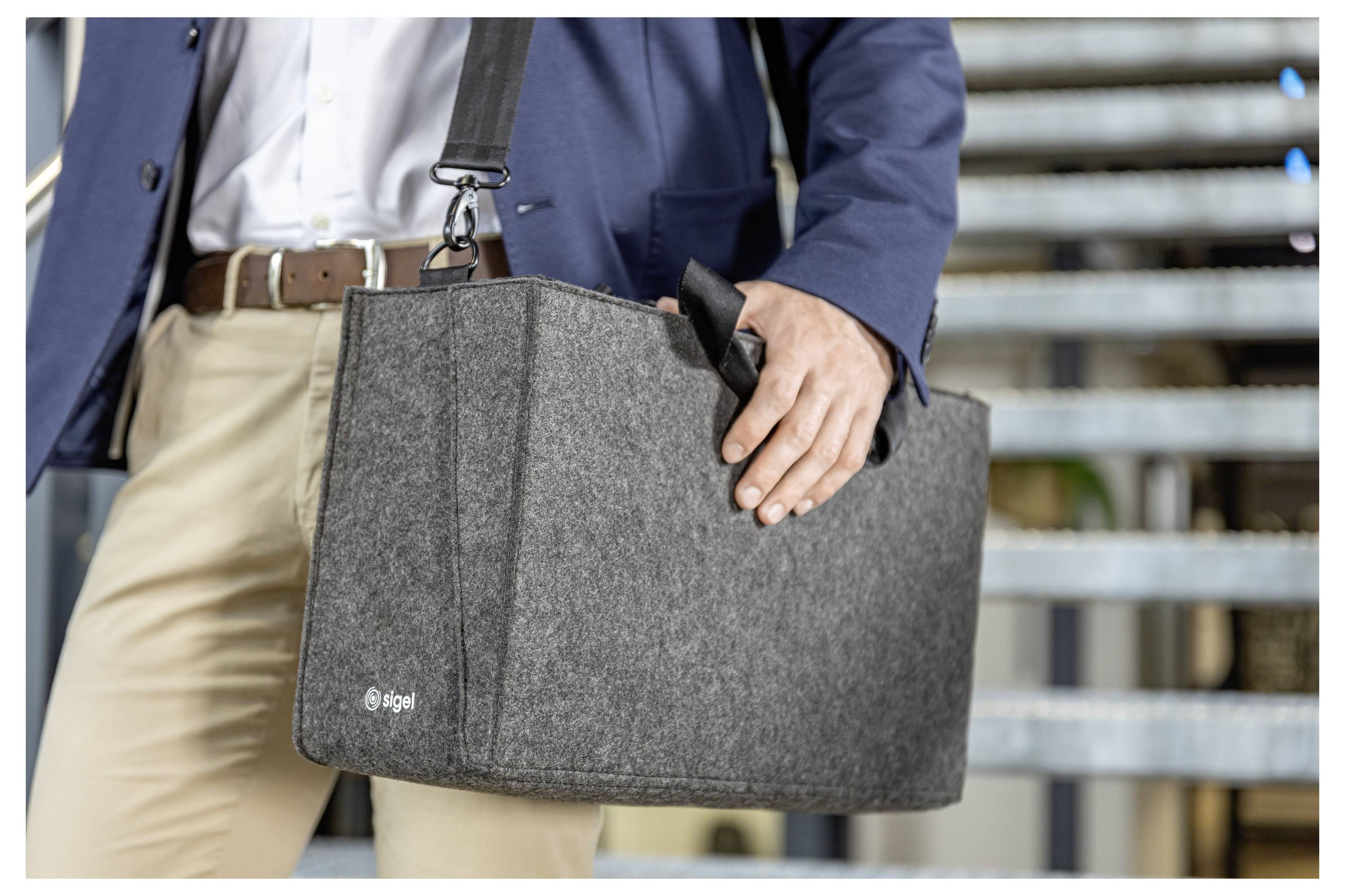 Nahaufnahme einer Person in einem Blazer, die eine graue Filz-Laptoptasche mit Schultergurt hält, im Freien in der Nähe von Metalltreppen.