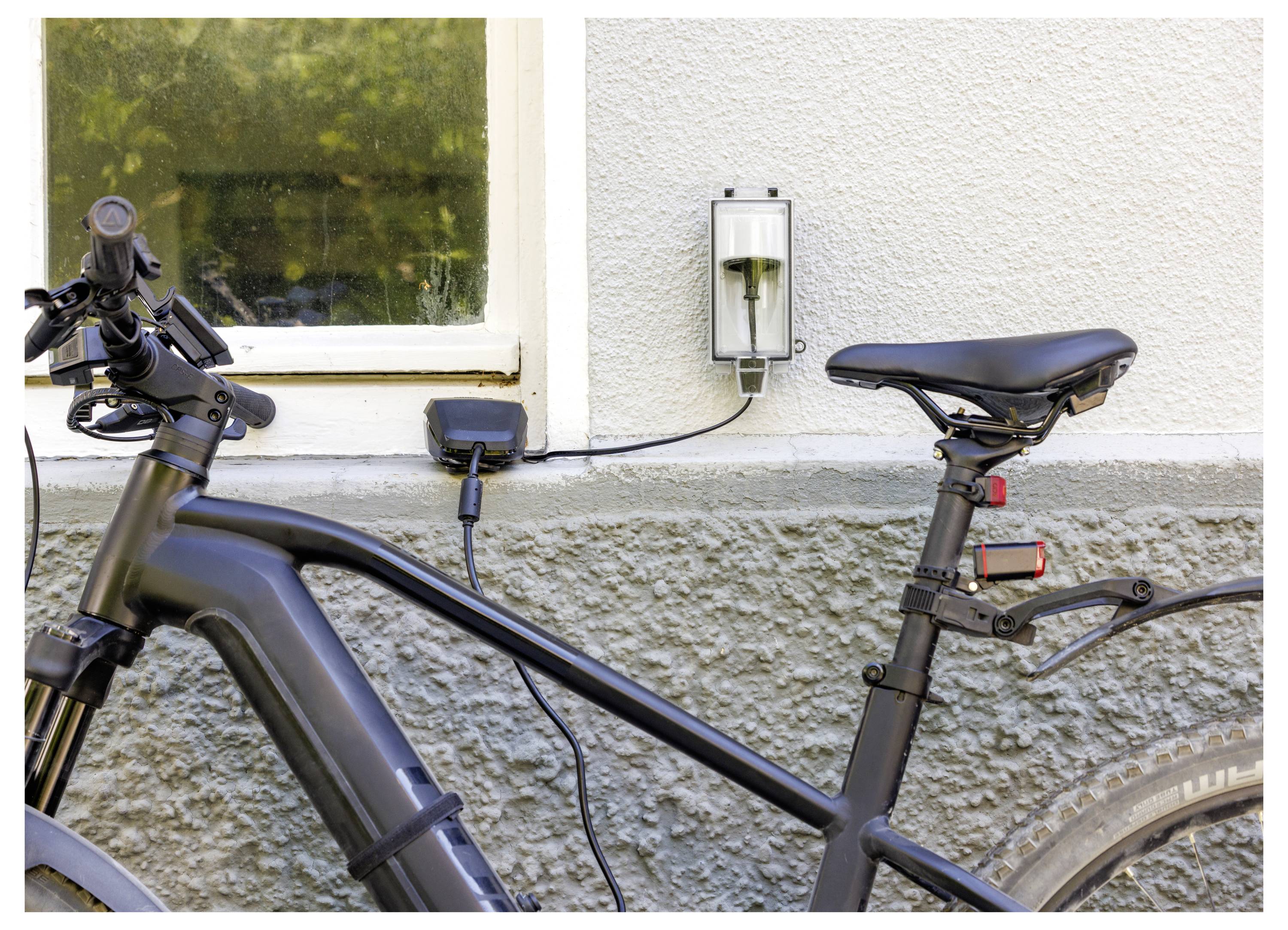 Elektrofahrrad, das an einem Außenwandladegerät neben einem Fenster angeschlossen ist und den Akku am Rahmen zeigt, der gerade aufgeladen wird.