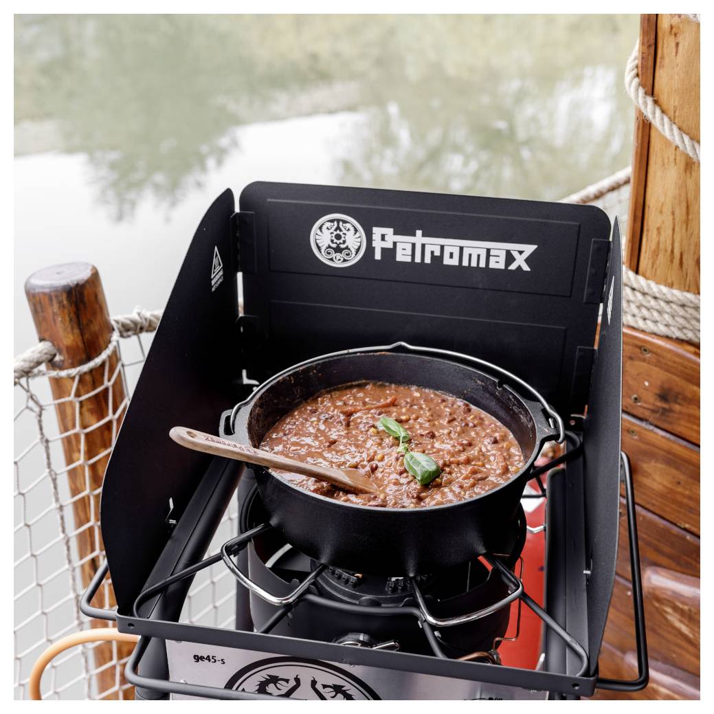 Ein gusseiserner Topf mit köchelndem Eintopf steht auf einem Petromax-Campingkocher im Freien am Wasser, mit einem Holzlöffel und einer Basilikum-Garnierung.
