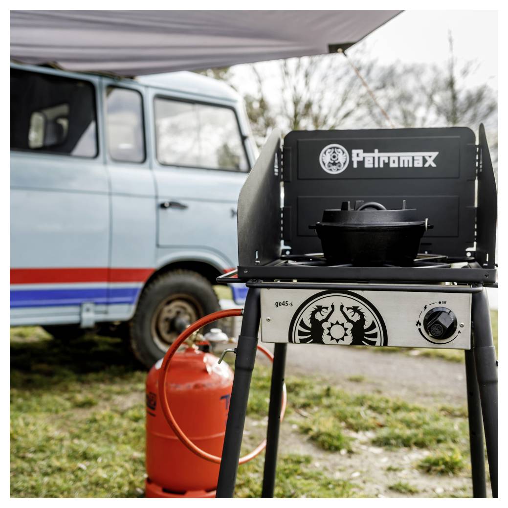 Tragbarer Petromax-Gaskochfeld mit einem Topf vor einem Vintage-Wagen, neben einer roten Propangasflasche unter einem Vordach im Freien.