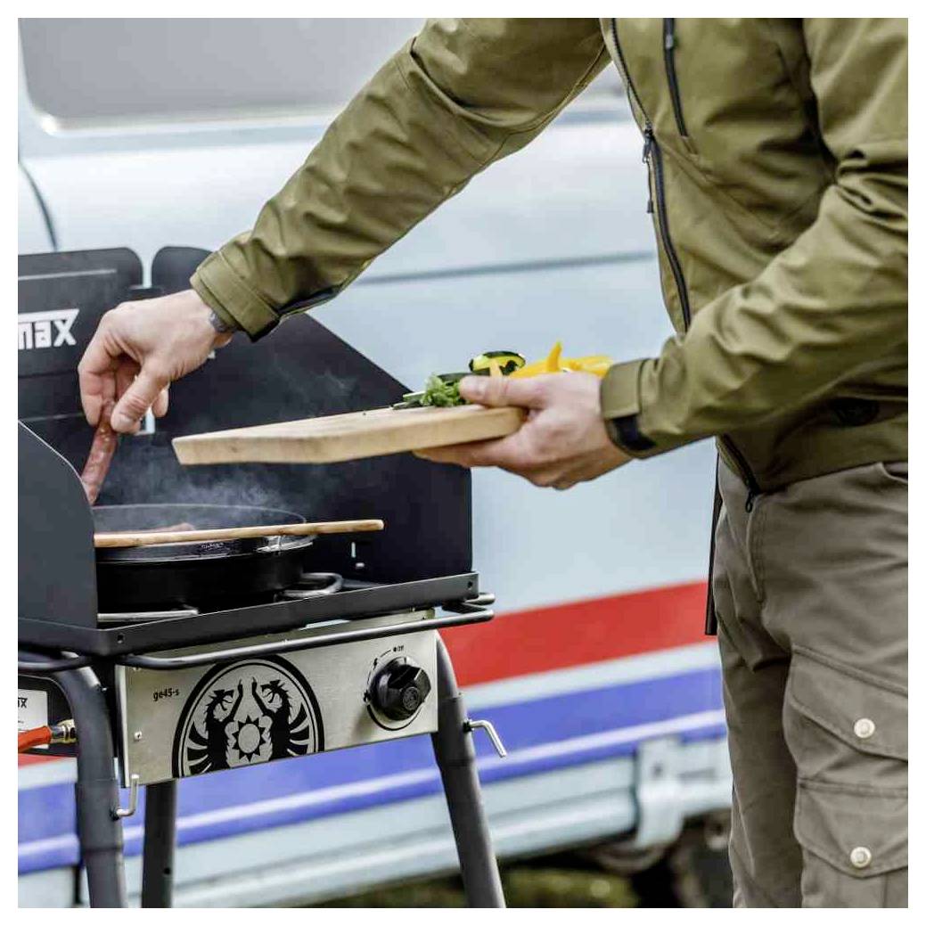 Person grillt Lebensmittel im Freien, hält ein Holzservierbrett neben einem tragbaren Barbecue-Grill mit Lebensmitteln, die darauf kochen.