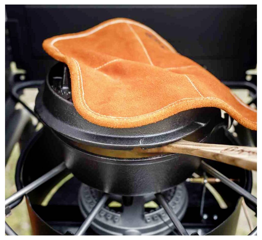 Orangener Ofenhandschuh hebt einen schwarzen Deckel auf einem Campingtopf über einem Herd an, mit einem sichtbaren Holzgriff.