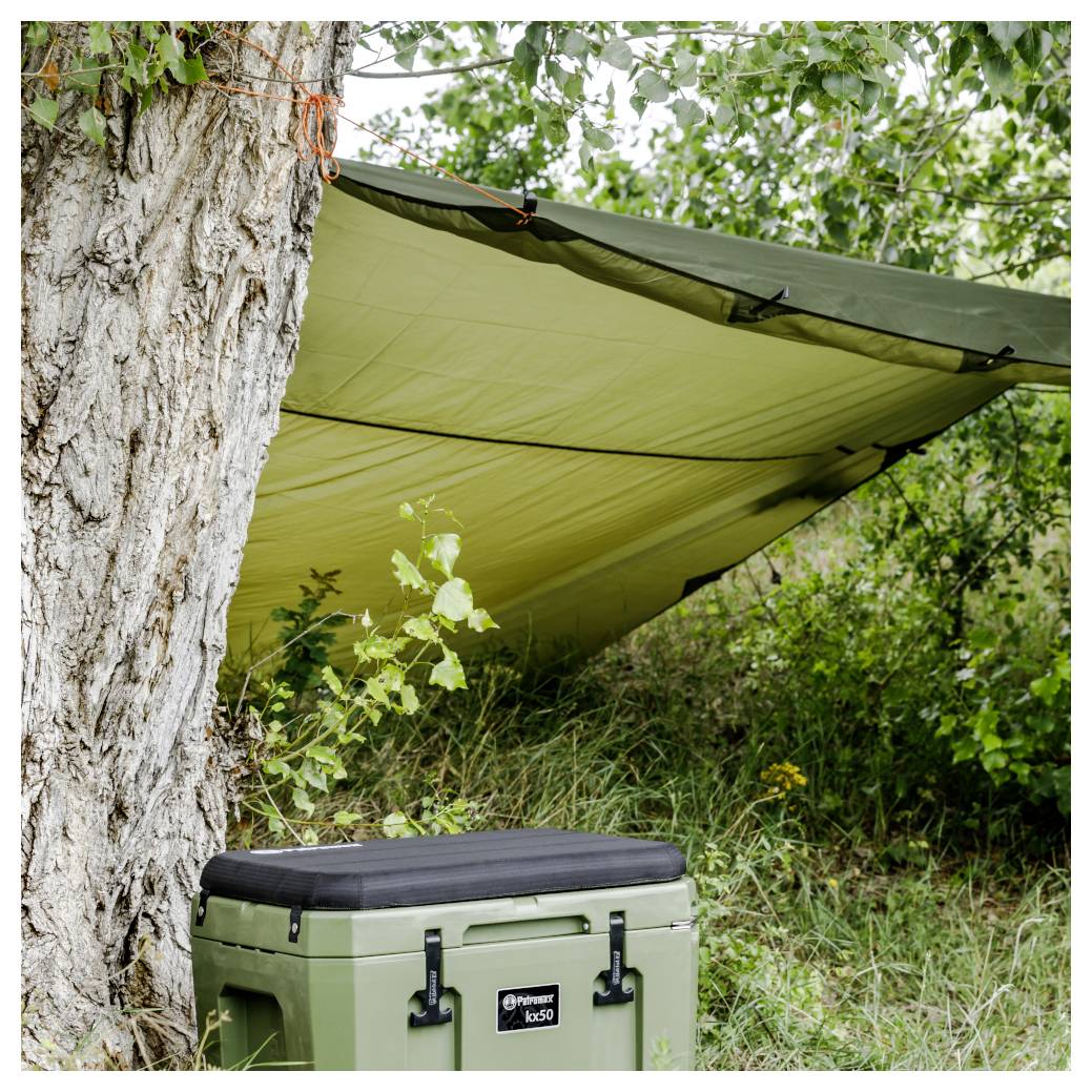 Camping-Zeltplane an einem Baum über einer olivefarbenen hartschaligen Kühlbox im hohen Gras und Gestrüpp befestigt.