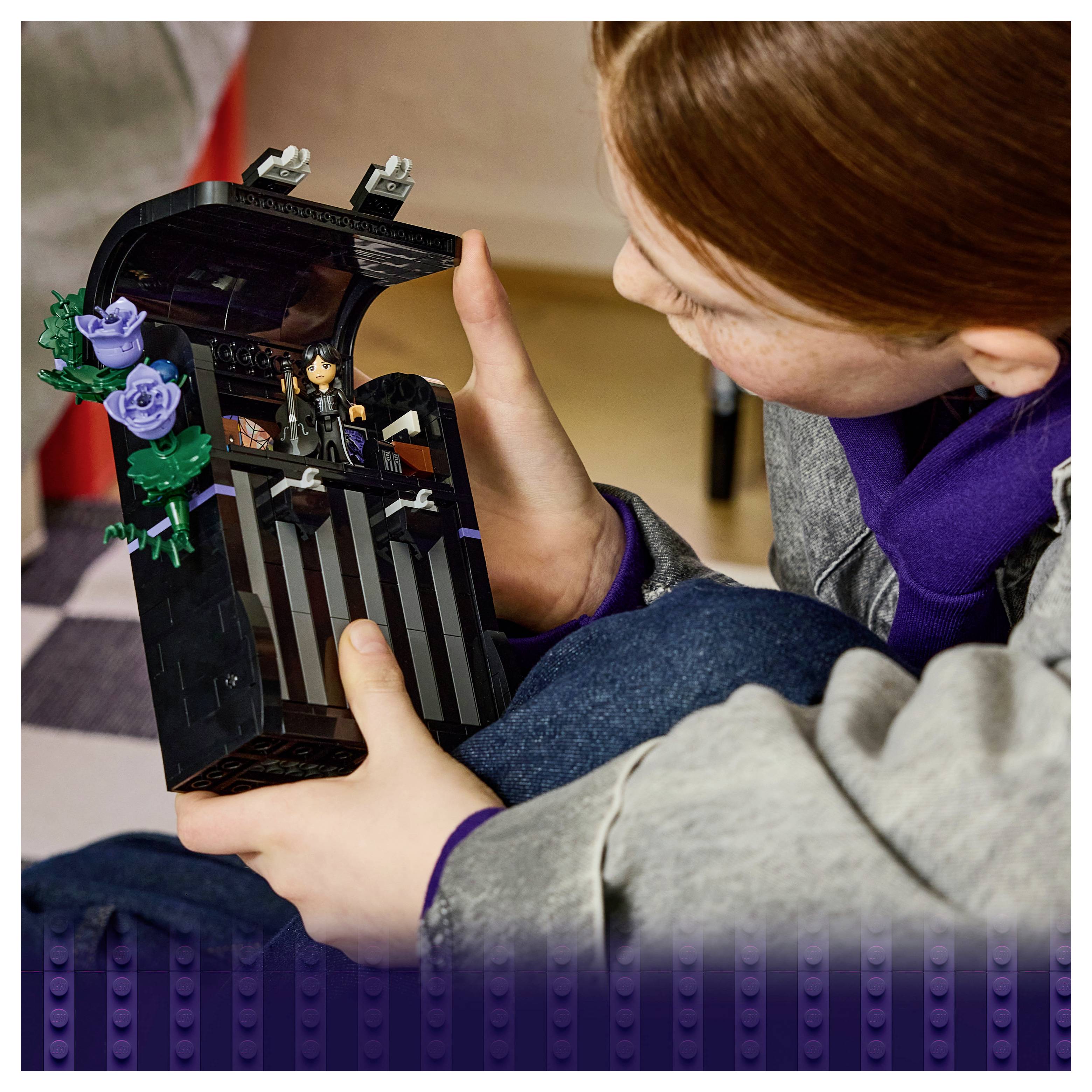 Ein Kind untersucht eine schwarze LEGO-Spielzeugtruhe oder einen Sarg, der mit violetten Rosen verziert ist und eine kleine Figur im Inneren enthält.