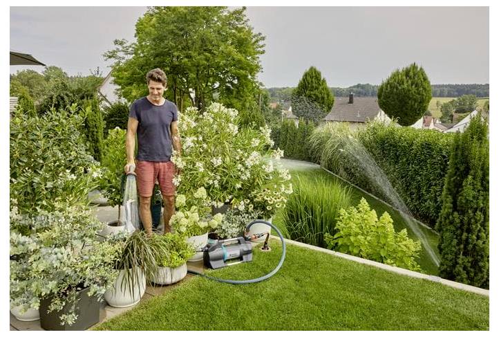 Ein Mann, der einen angelegten Garten mit einem Schlauch und einem Sprinkler neben Topfpflanzen bewässert, mit Hecken und Häusern im Hintergrund.