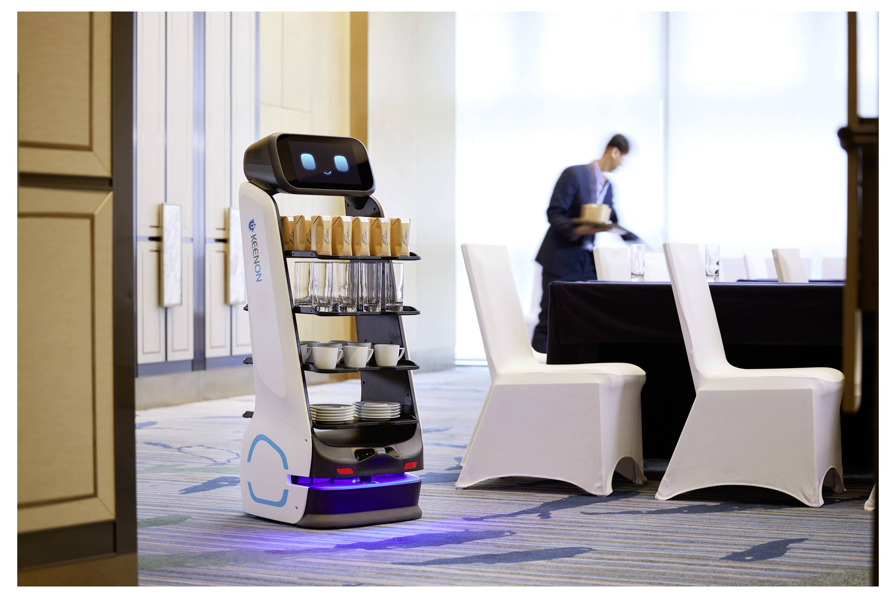 Autonomer Serviceroboter, der Tabletts und Tassen in einem Konferenzraum transportiert, während eine Person im Hintergrund Tischdekorationen arrangiert.