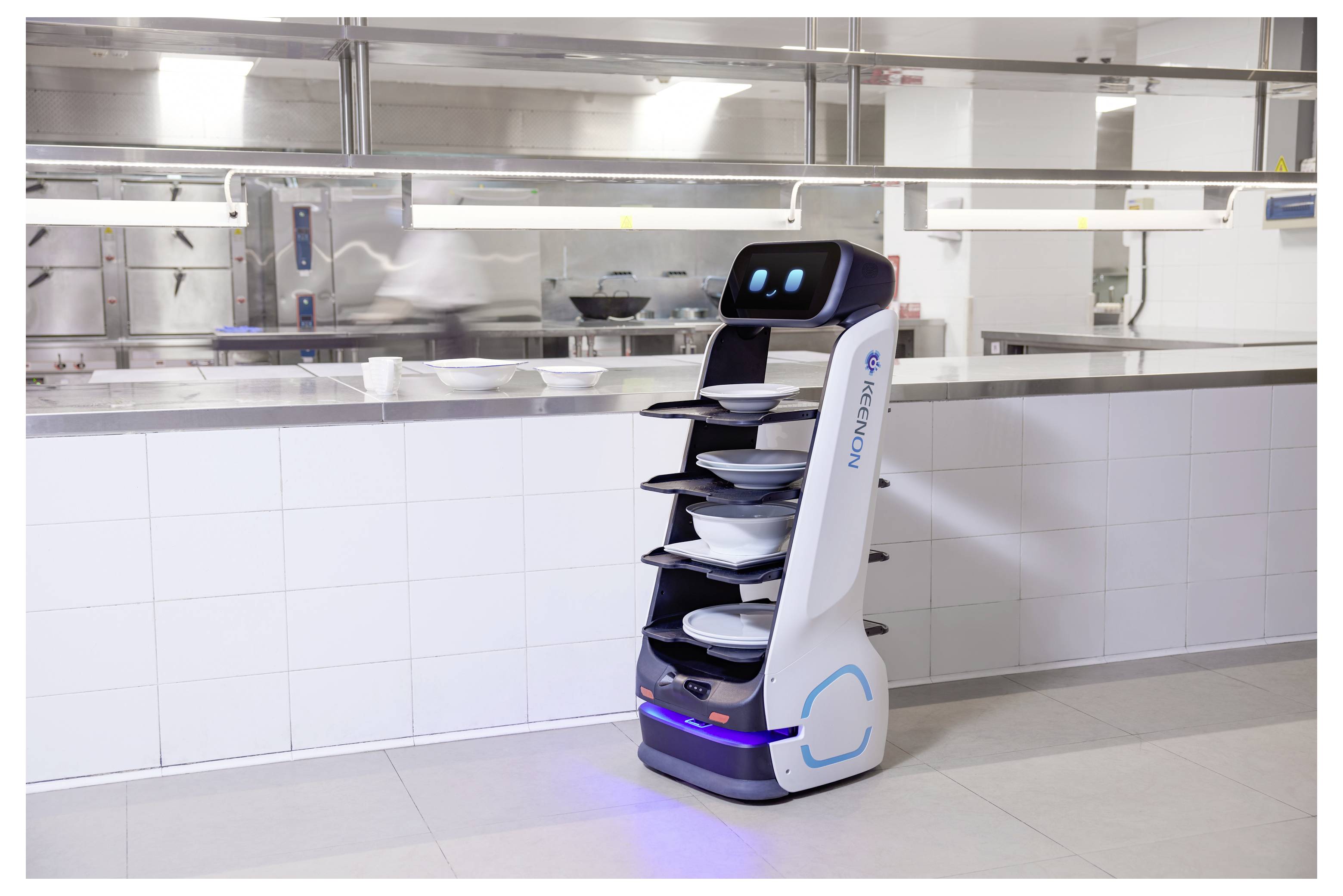 Autonomer Serviceroboter mit gestapelten Tabletts steht in einer hellen kommerziellen Küche oder Cafeteria-Servicebereich.