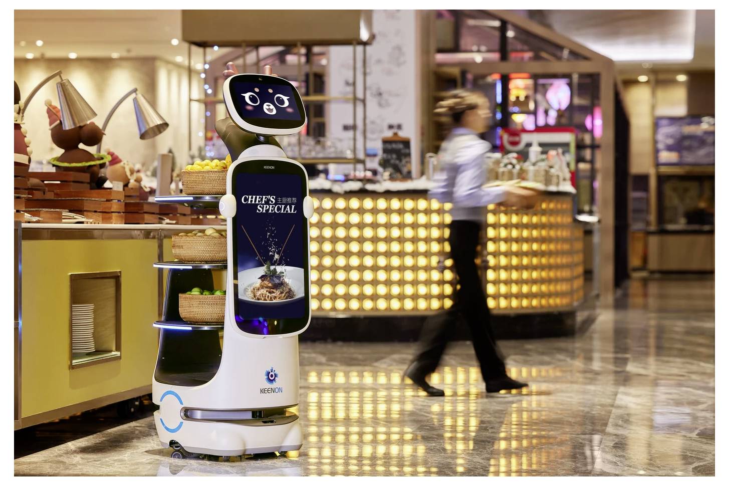 Ein Restaurantservice-Roboter mit einem Schild „SPEZIALITÄT DES CHEFS" steht in der Nähe eines Buffettresen, während eine unscharfe Person in einem Einkaufszentrum oder einer Foodcourt vorbeigehen.