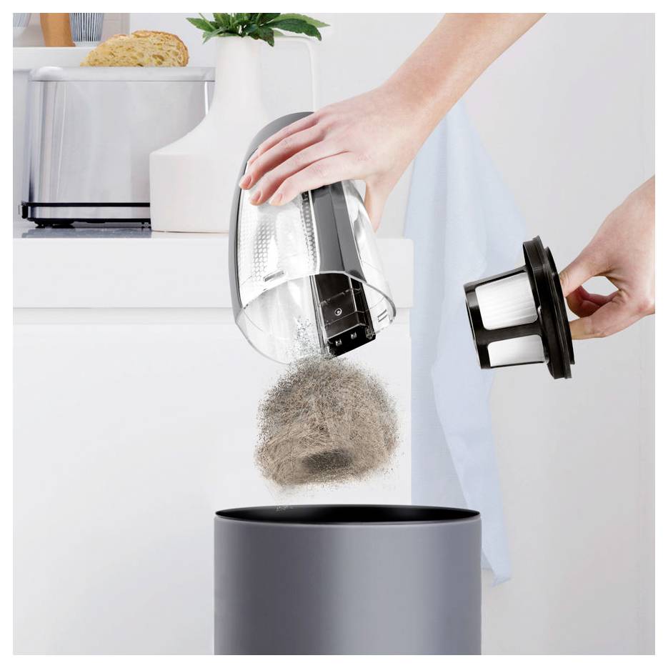 Person, die Staub aus einem Stabstaubsauger in einen Mülleimer leert, während sie den Filter in einer hellen Küche entfernt.