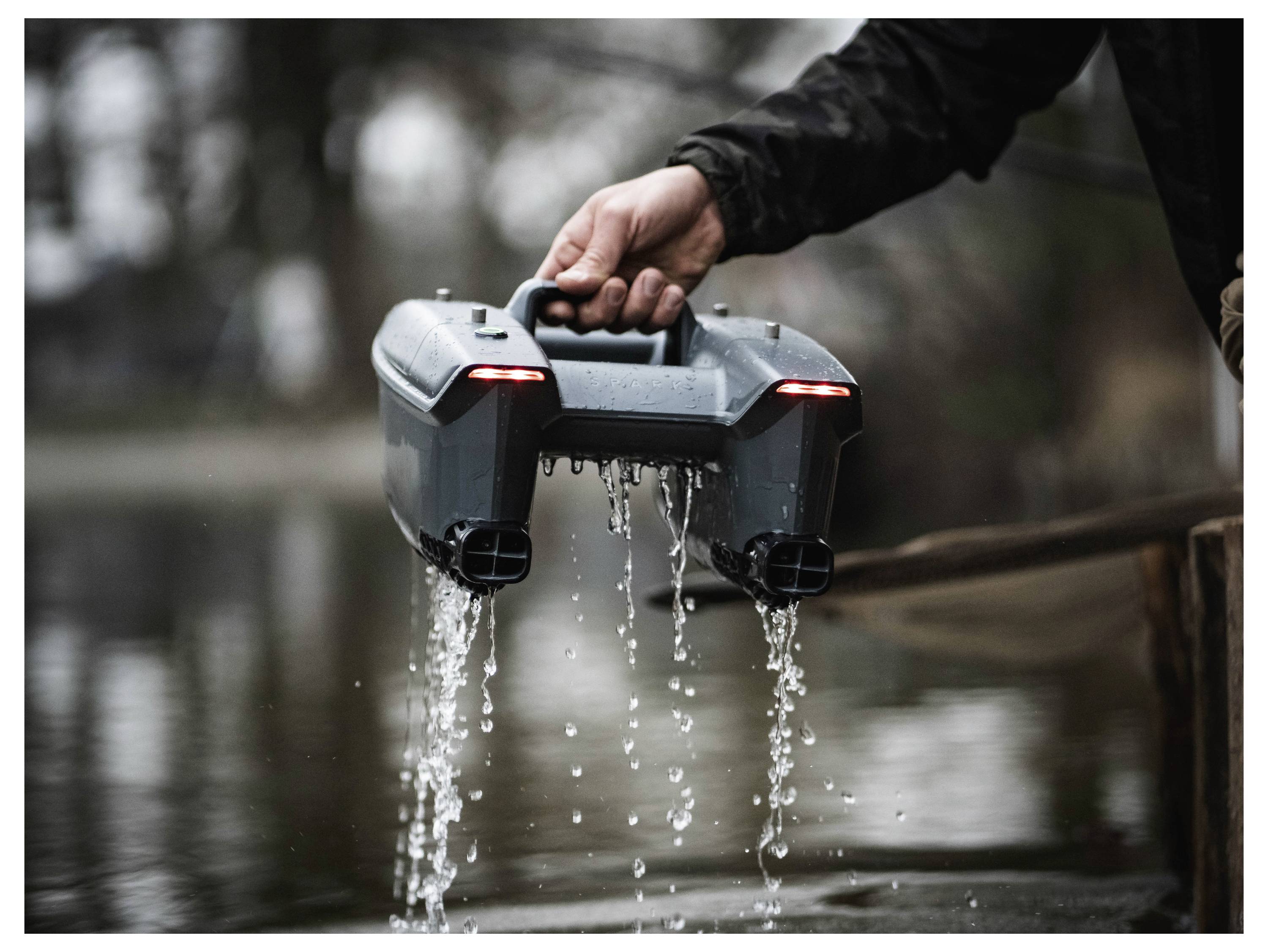 Eine Person hält ein graues tragbares Gerät an seinem Griff, während Wasser aus zwei vorderen Öffnungen auf überflutetes Gelände fließt.