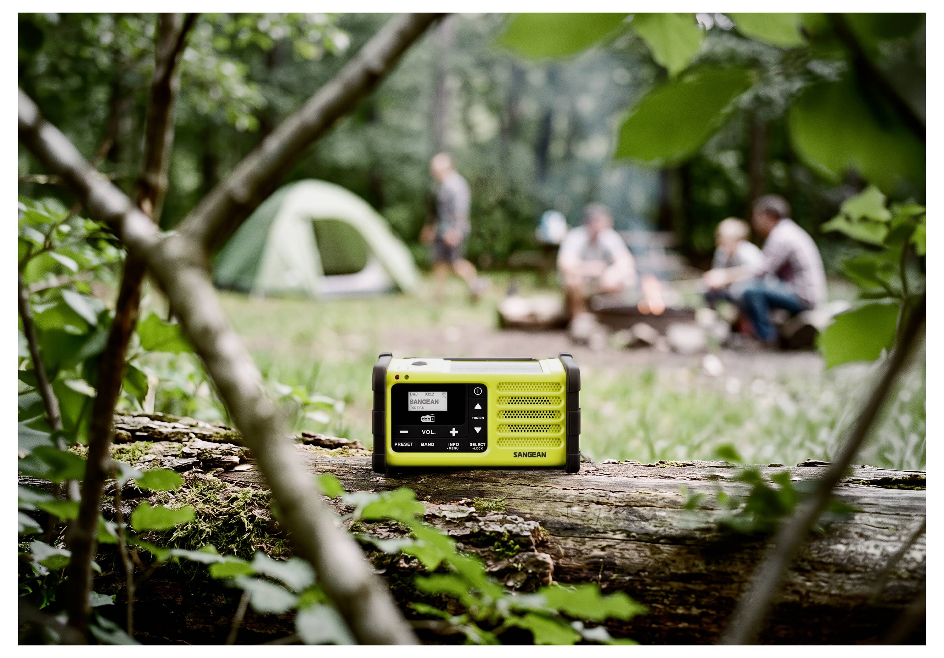 Tragbares gelbes Radio auf einem gefallenen Baumstamm auf einem Campingplatz, mit unscharfen Menschen und Lagerfeuer im Hintergrund.