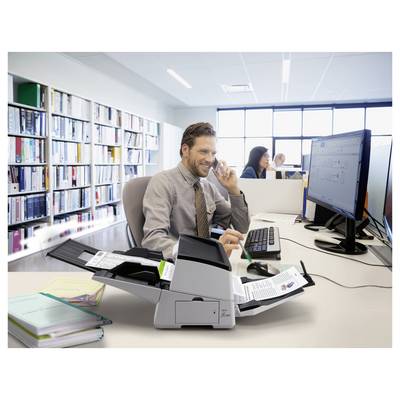 Mann arbeitet an einem Schreibtisch in einem Büro mit einem Telefon und einem Computer neben einem Drucker/Scanner, mit Kollegen im Hintergrund.