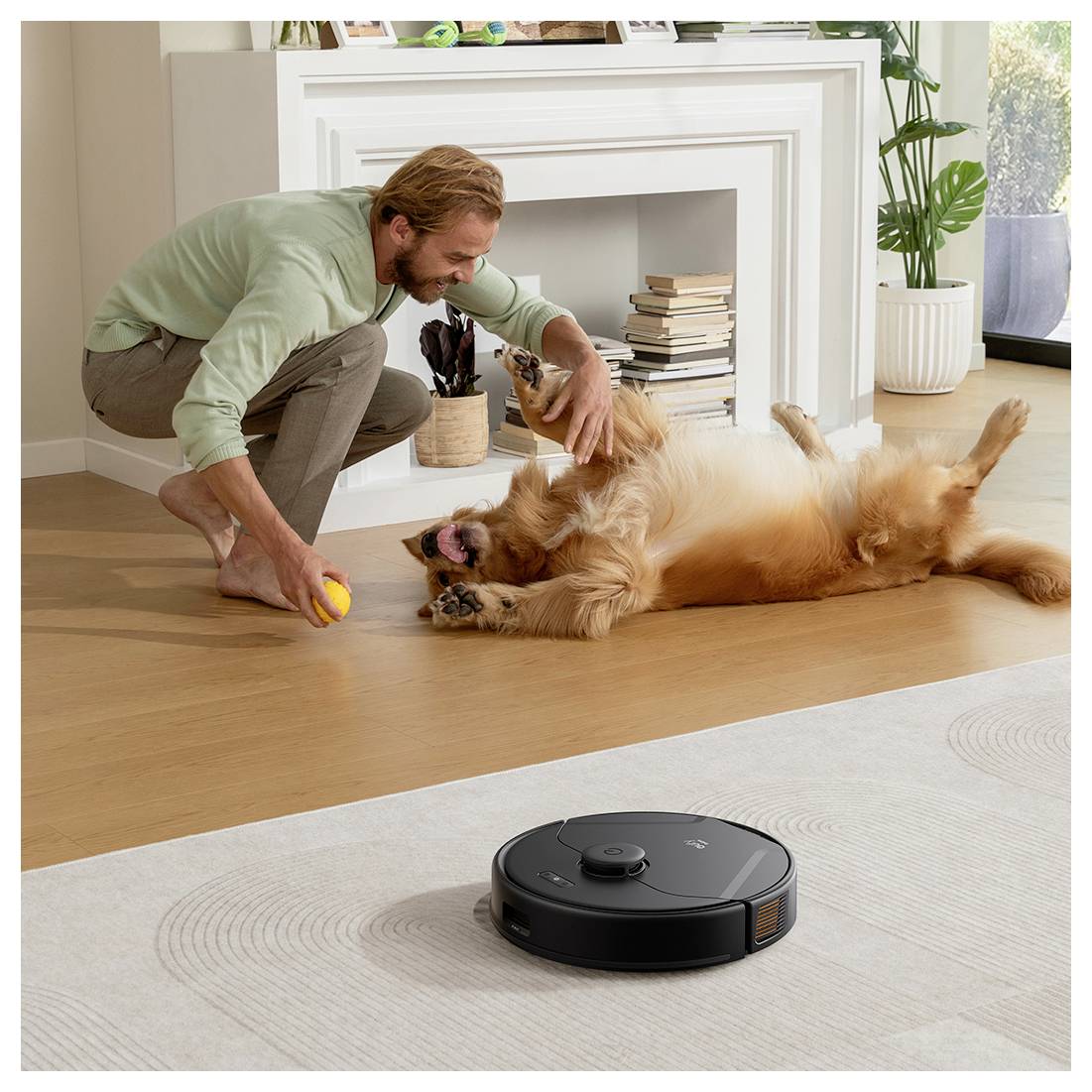 Ein Mann spielt mit einem Golden Retriever in einem hellen Wohnzimmer, während ein schwarzer Roboterstaubsauger den Teppich im Vordergrund reinigt.