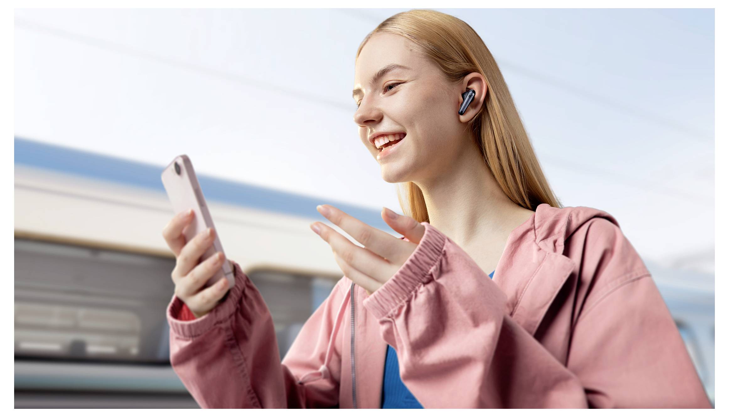 Lächelnde Frau in einer rosa Jacke mit kabellosen Ohrhörern, die auf ihr Telefon schaut und gestikuliert, als würde sie einen Videoanruf führen.