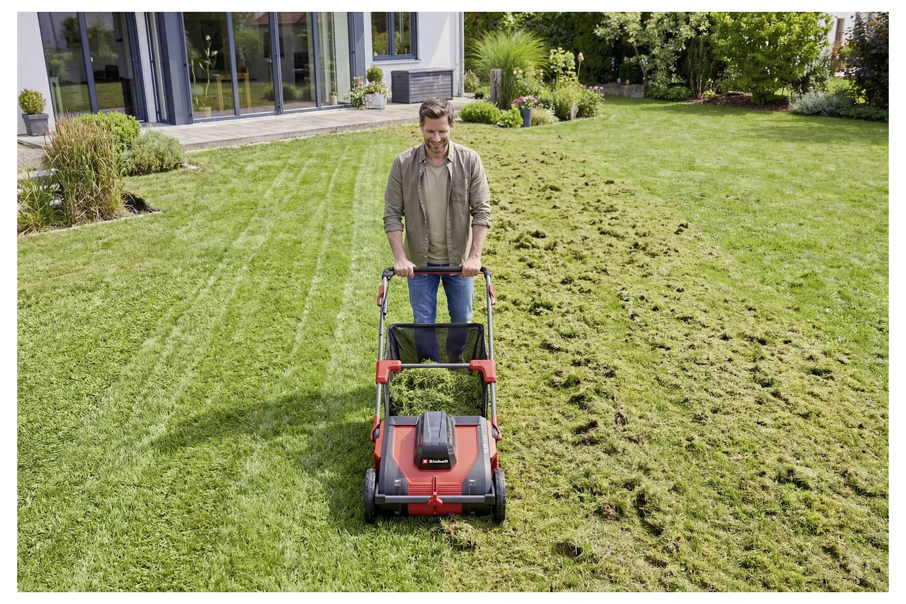 Ein Mann mäht einen Rasen mit einem roten Rasenmäher und zeigt dabei ein klares Streifenmuster neben einem ungemähten Bereich in einem gestalteten Garten.