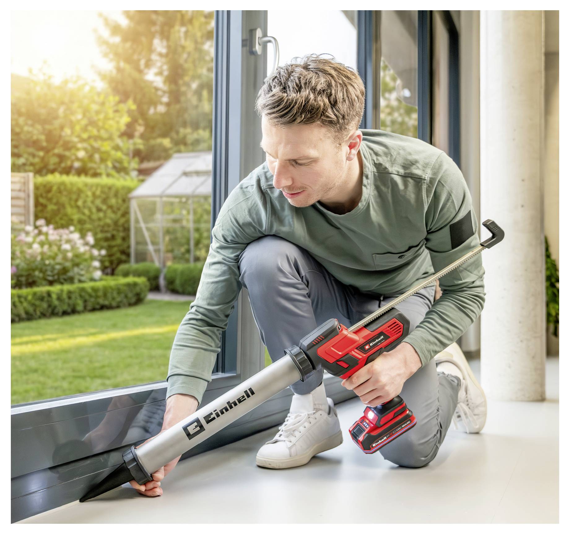 Ein Mann kniet drinnen und nutzt einen kabellosen Laubblaser, um Schmutz in der Nähe einer Glasschiebetür zu beseitigen, während ein sonniger Garten draußen sichtbar ist.