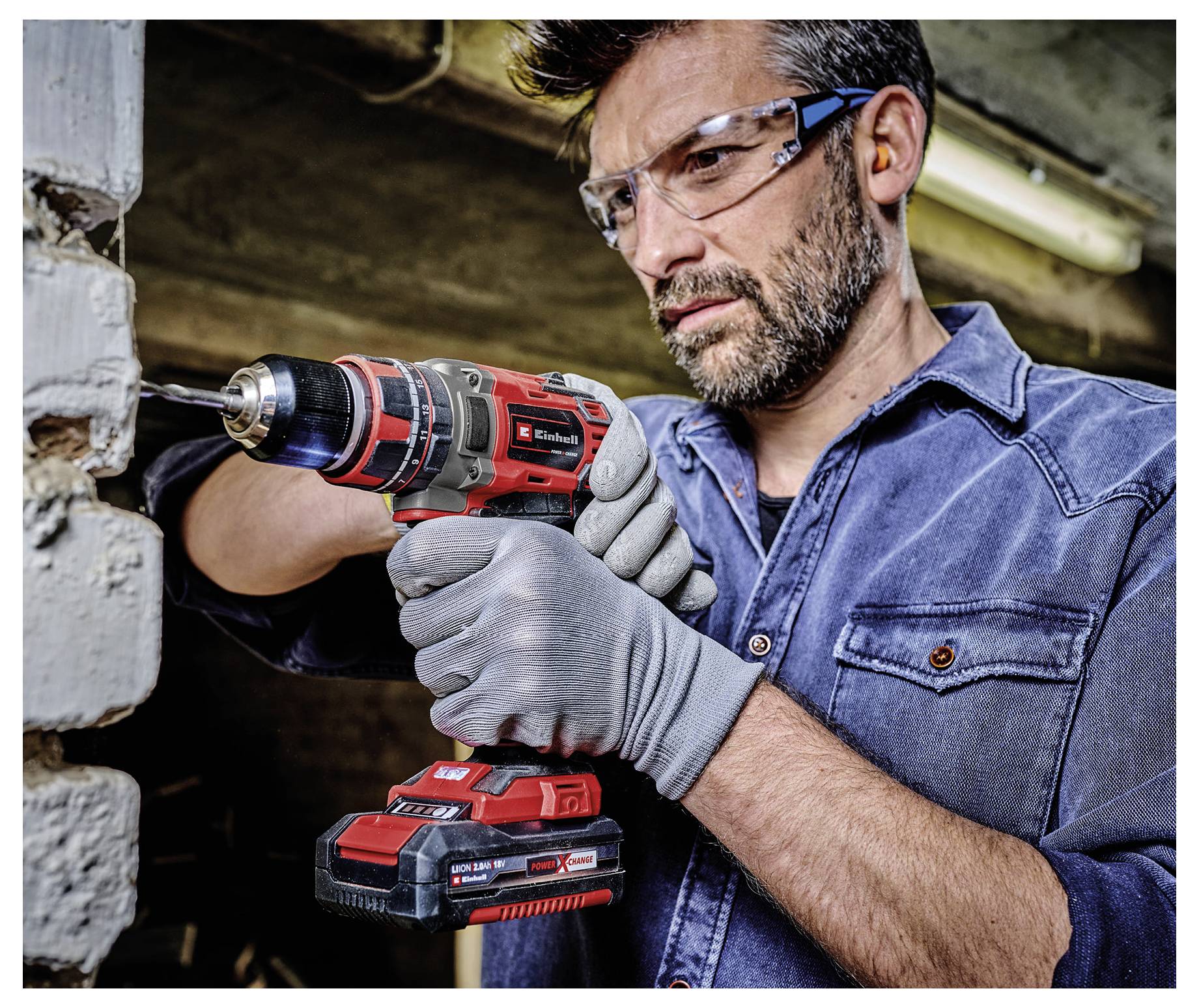 Ein Mann, der einen kabellosen Bohrer verwendet, um in eine Betonwand zu bohren, trägt dabei eine Schutzbrille und Handschuhe in einer Werkstatt oder auf einer Baustelle.