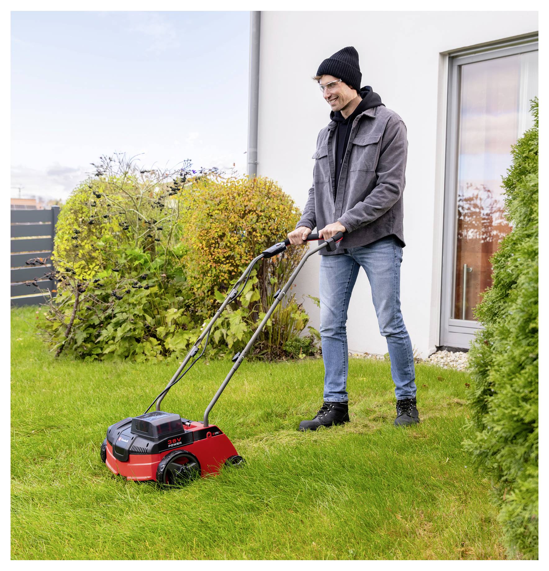 Ein Mann mäht einen Rasen mit einem roten Rasenmäher neben einem Haus und Büschen auf einem Grasgarten.