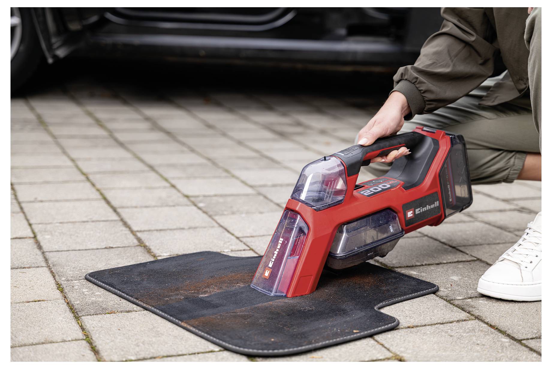 Eine Person benutzt einen roten tragbaren Staubsauger, um eine schwarze Autofußmatte auf einer gepflasterten Auffahrt neben einem geparkten Fahrzeug zu reinigen.
