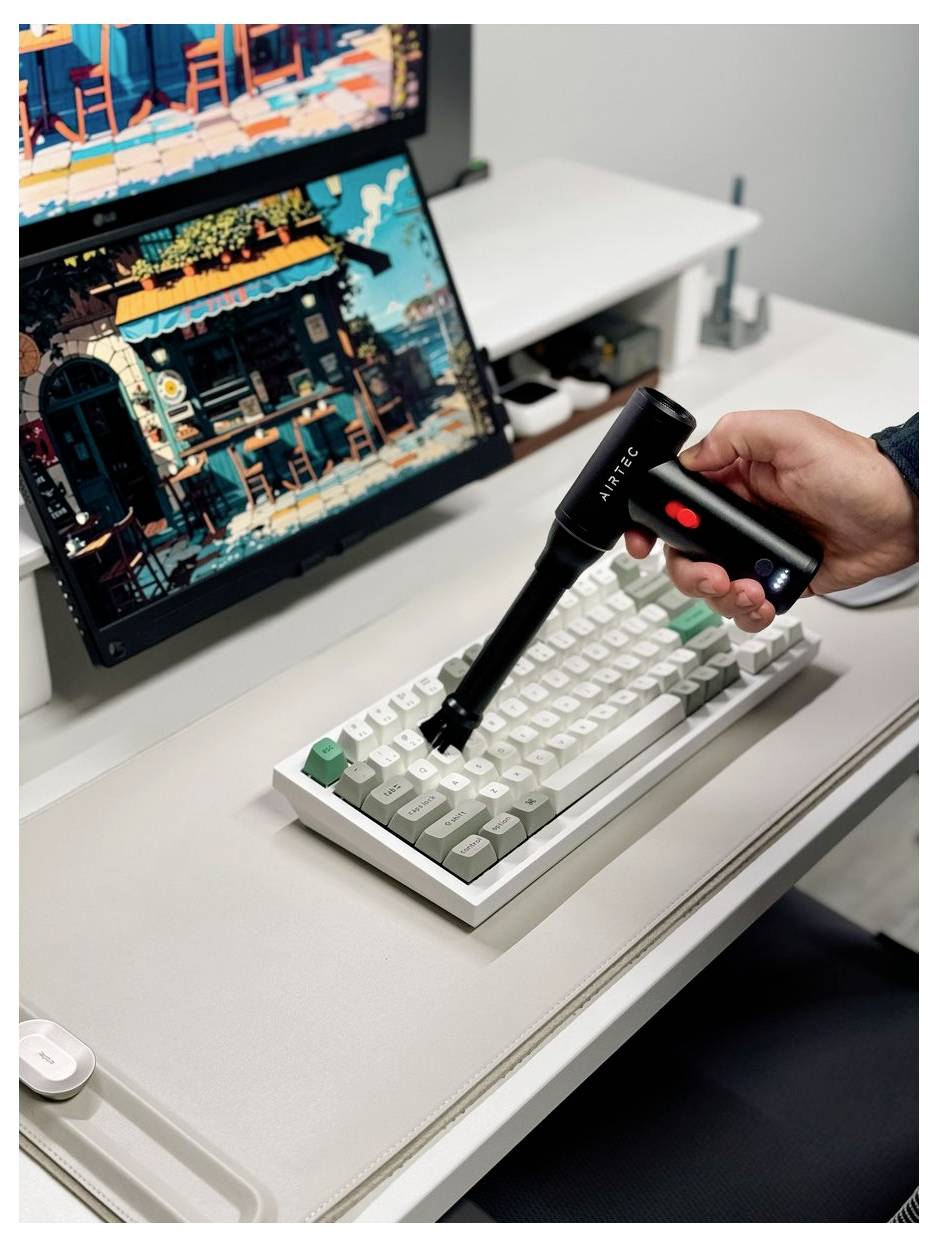 Tragbarer Druckluft-Reiniger zum Reinigen einer kompakten mechanischen Tastatur auf einem Schreibtisch, mit einem Monitor, der ein farbenfroher Pixel-Art-Spiel im Hintergrund zeigt.
