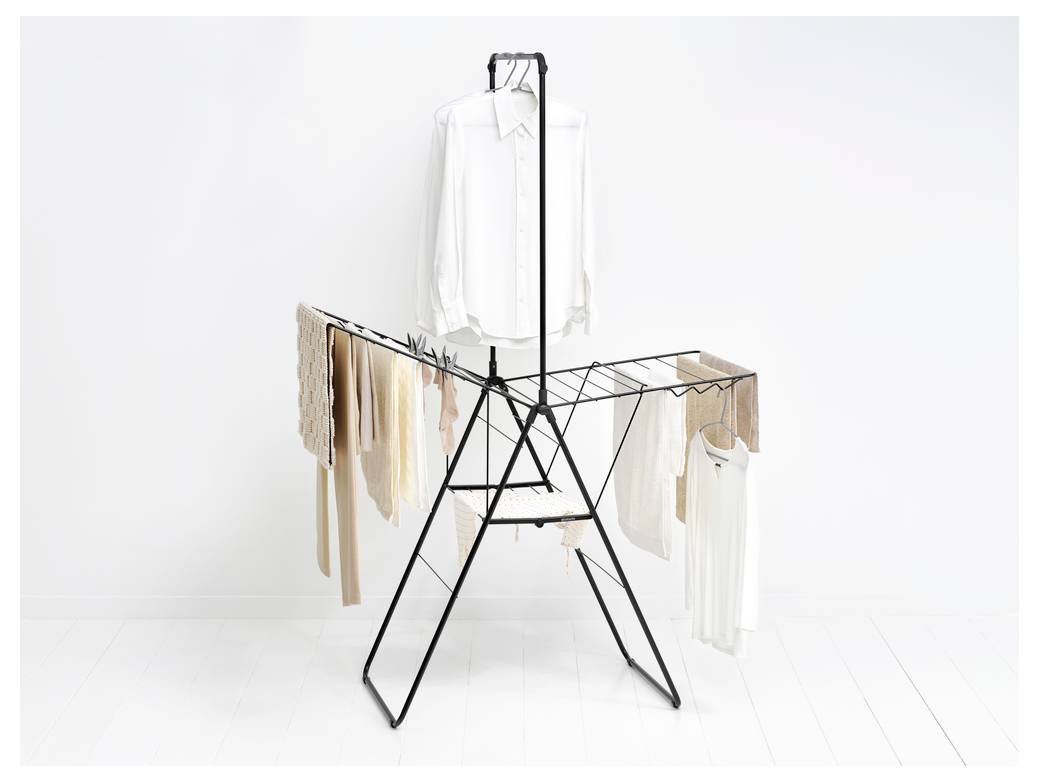 Ein schwarzes Metallwäschegestell in einem weißen Raum hält hängende weiße Hemden und hellfarbige Kleidungsstücke, mit Handtüchern und Kleidung, die an den Seiten drapiert sind.