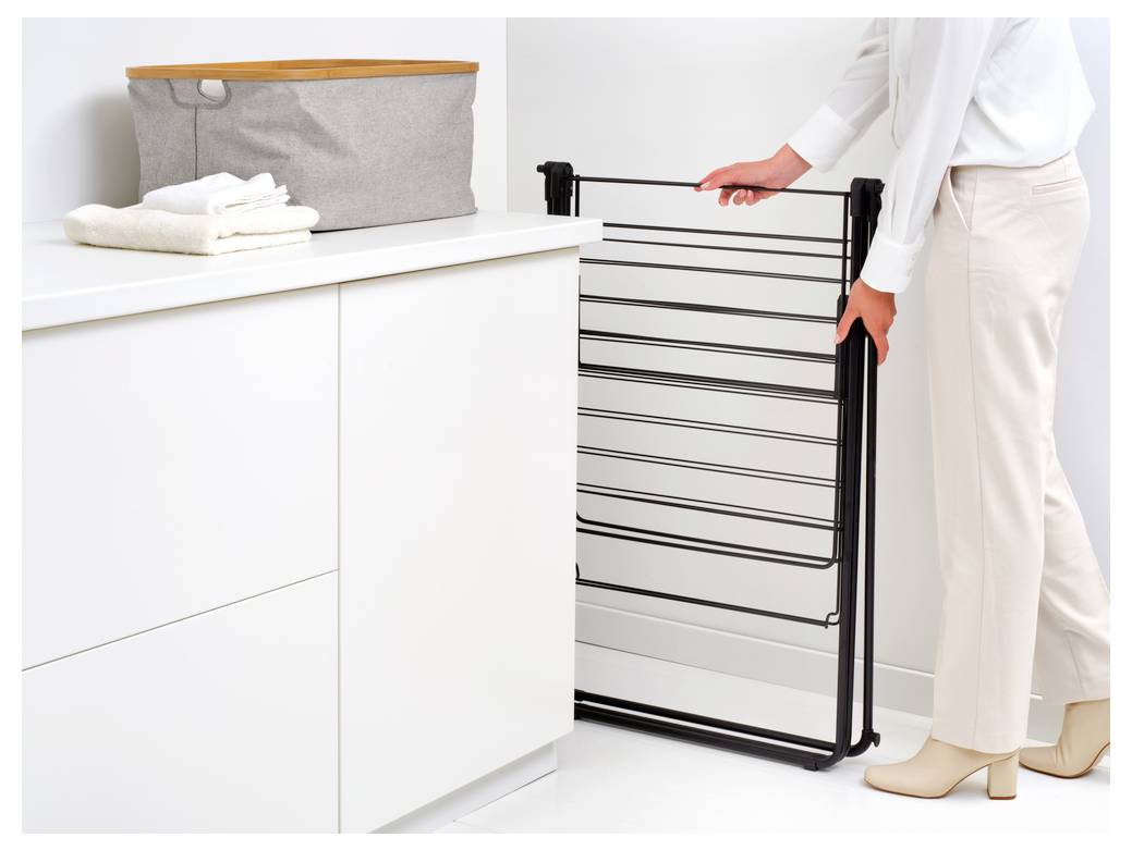 Eine Person faltet ein schwarzes Trockengestell neben einem weißen Schrank in einem hellen Waschraum.