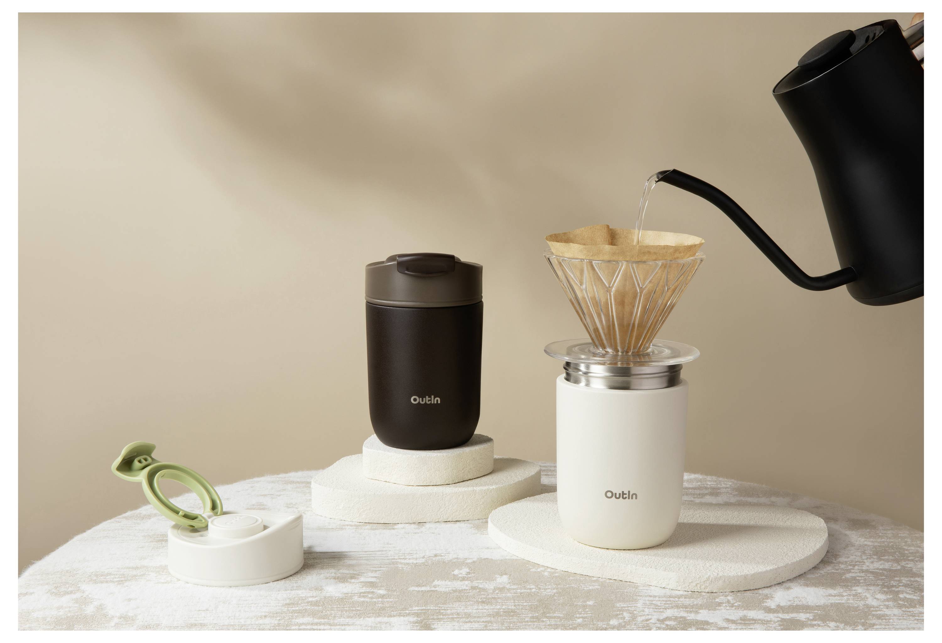 Minimalistische Kaffee-Ausstattung mit einem schwarzen Schwanenhals-Wasserkocher, der heißes Wasser in einen Pour-Over-Dripper auf einer weißen Tasse gießt, daneben ein schwarzer Reisebecher und ein grünes Clip-Werkzeug.