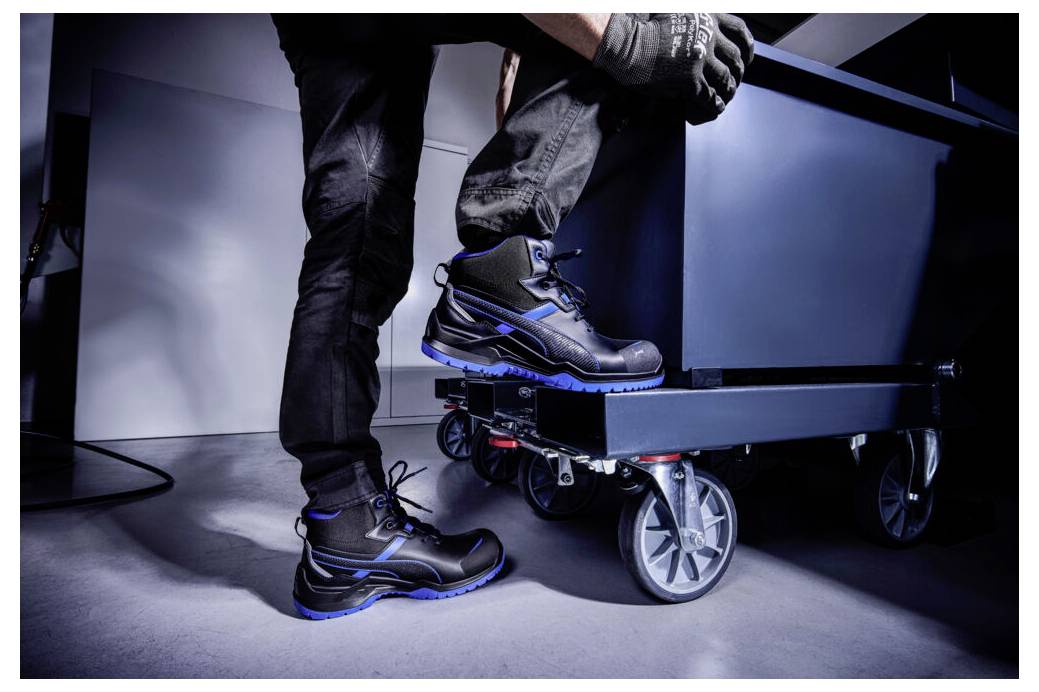 Nahaufnahme einer Person, die schwarze Arbeitsstiefel mit blauen Akzenten trägt und auf einen niedrigen Wagen oder ein niedriges Podest in einer studioähnlichen Umgebung steigt.