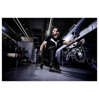 Mechaniker in schwarzer Arbeitskleidung hockt neben einem Fahrzeug in einer Werkstatt und inspiziert die Ausrüstung unter heller industrieller Beleuchtung.