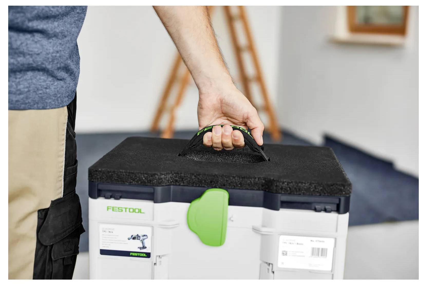Person carrying a Festool tool case by its handle in a workshop or room interior.<br><br>Eine Person trägt einen Festool-Werkzeugkoffer an seinem Griff in einer Werkstatt oder einem Innenraum.