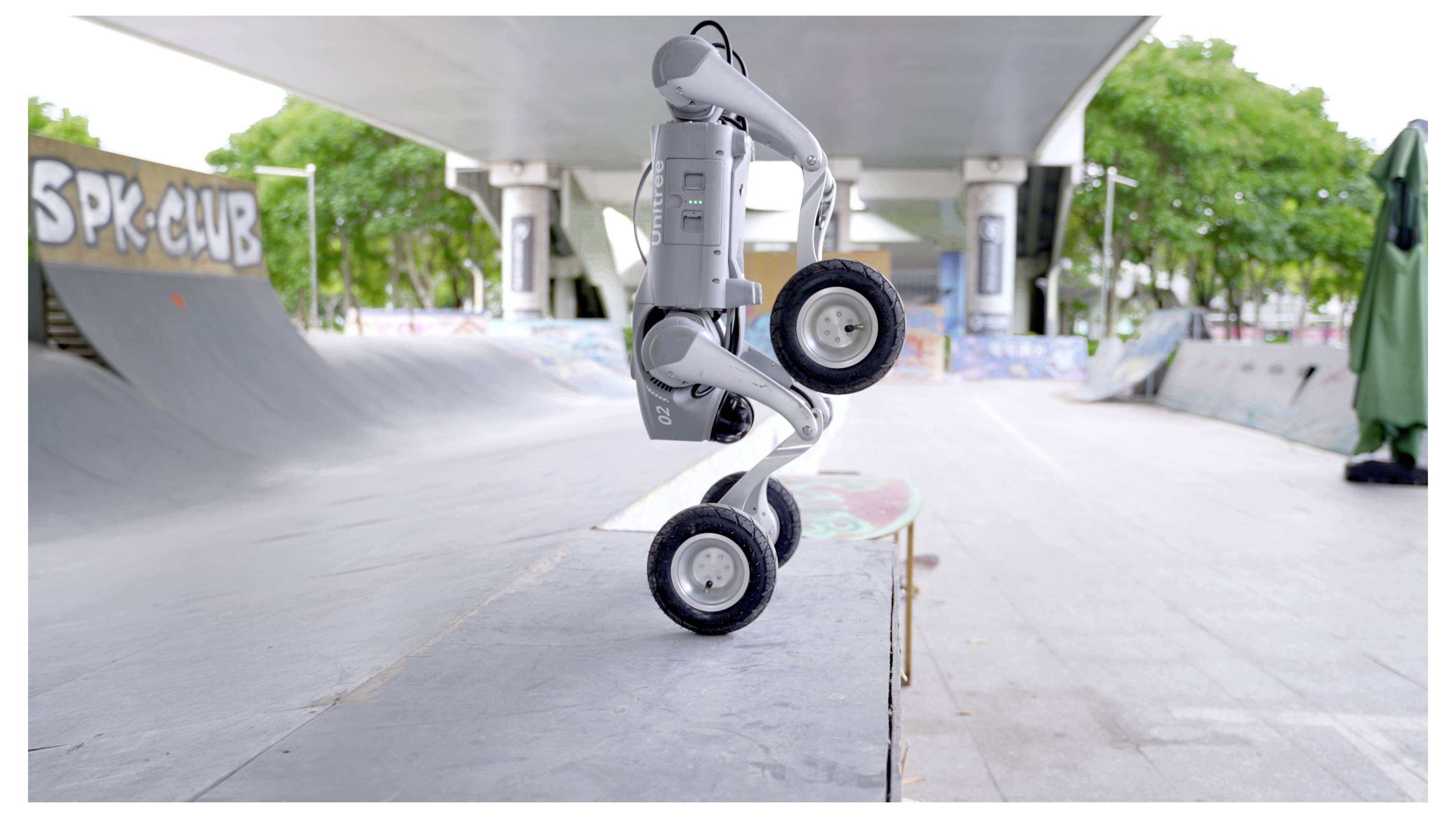 Ein kleiner grauer Roboter balanciert auf zwei Rädern in einem Skatepark unter einer Betonüberführung, mit Rampen und Graffiti im Hintergrund.