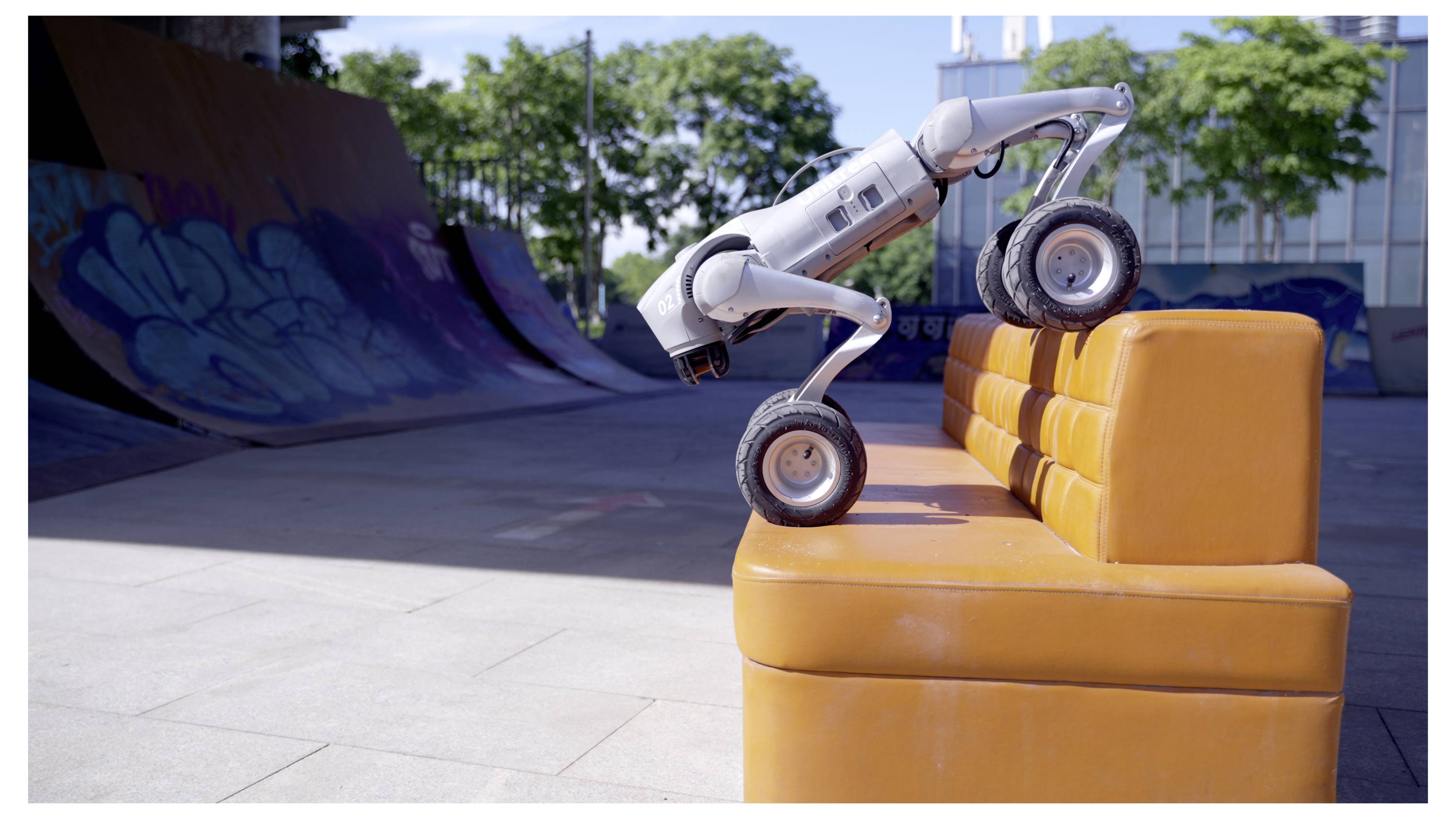 Ein kleiner Roboter mit Rädern erklimmt orangefarbene Schaumstoffstufen in einem Outdoor-Skatepark, wobei eine Graffiti-Rampe im Hintergrund zu sehen ist.