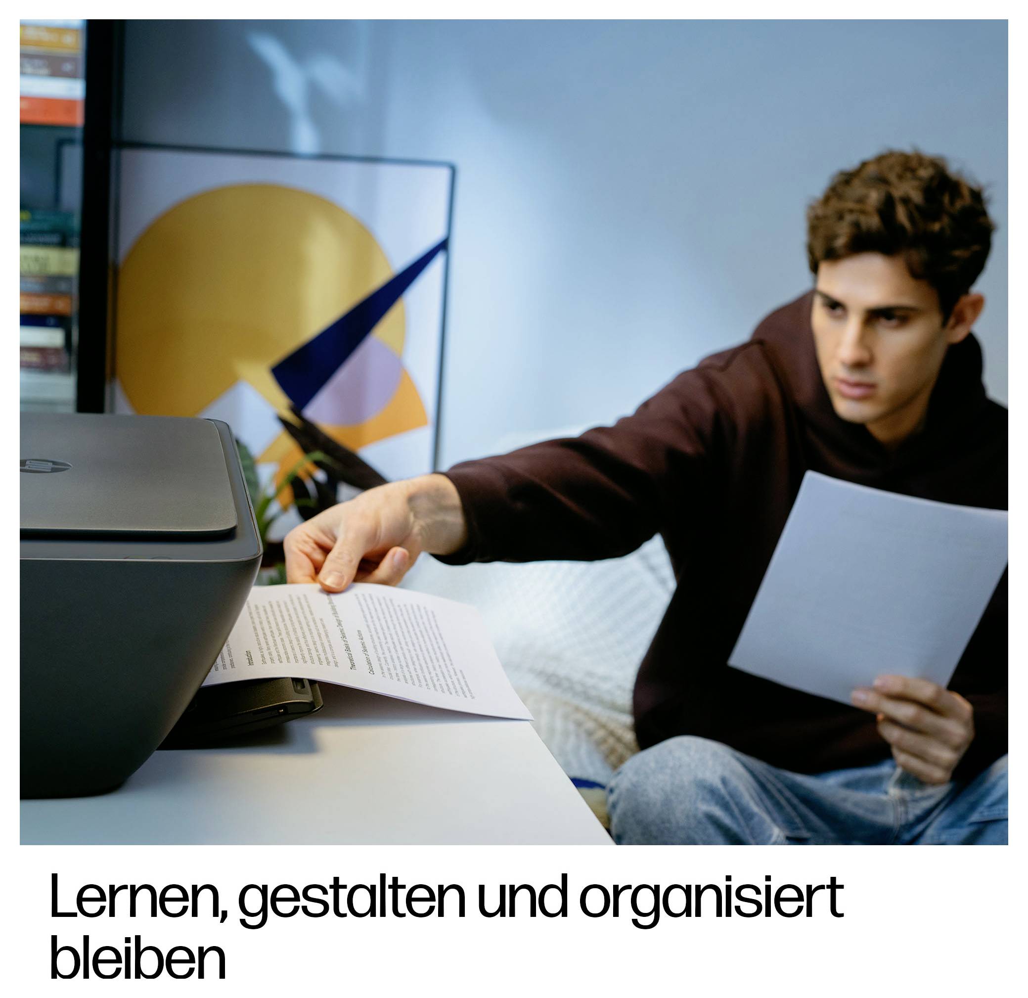 Junger Mann an einem Schreibtisch greift nach einer gedruckten Seite, während er eine andere hält, neben einem Drucker; deutscher Text liest sich „Lernen, gestalten und organisiert bleiben".
