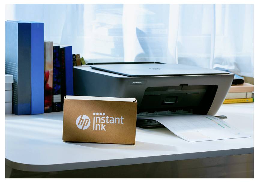 HP-Drucker auf einem Schreibtisch mit einer "HP Instant Ink"-Box davor, Papier in das Fach eingelegt, und Bücher neben einem Fenster