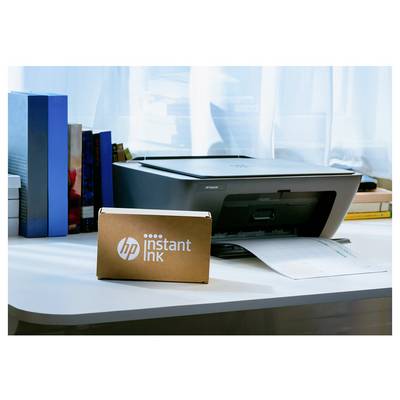 HP-Drucker auf einem Schreibtisch mit einer "HP Instant Ink"-Box davor, Papier in das Fach eingelegt, und Bücher neben einem Fenster