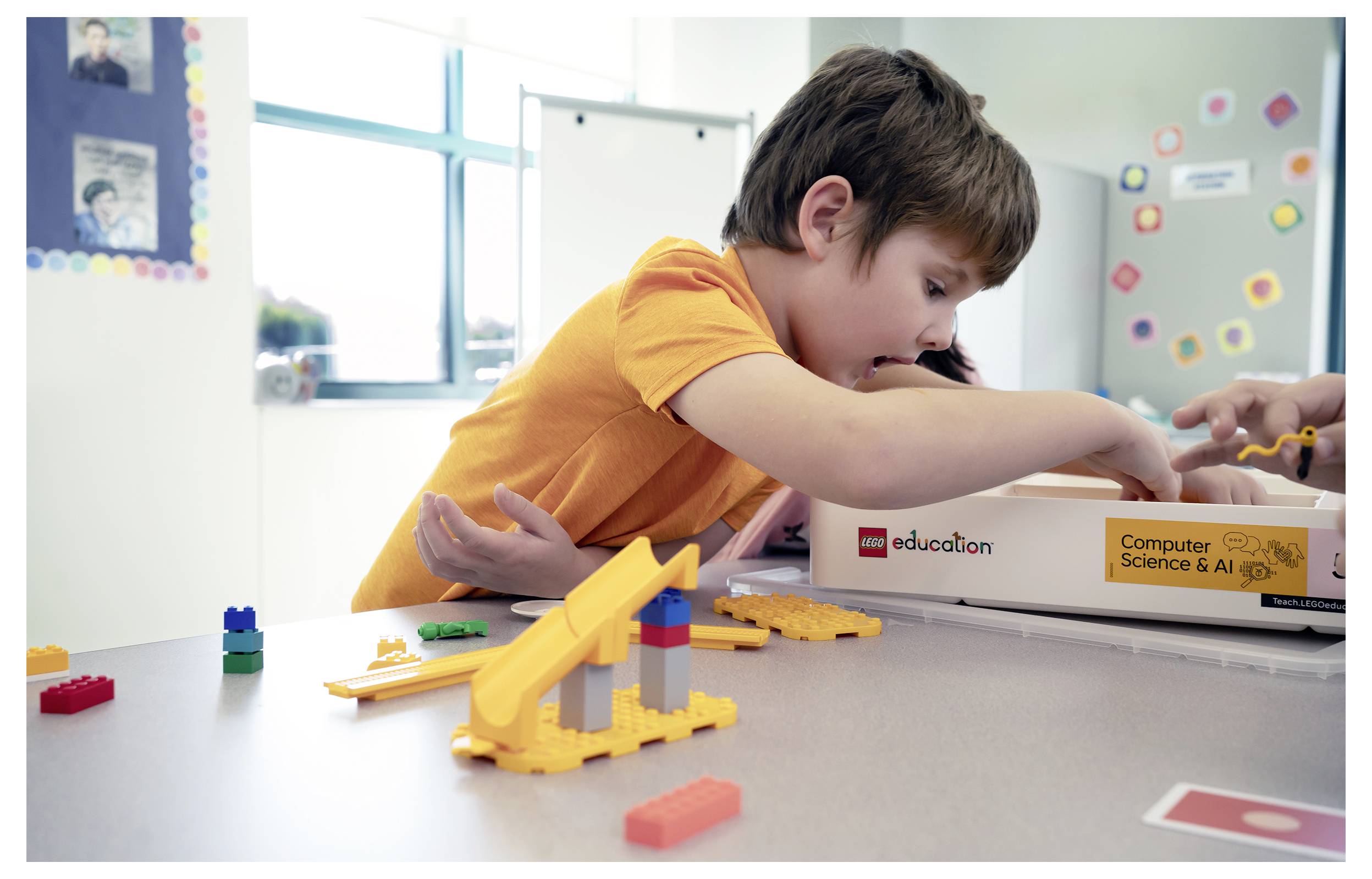 Junger Junge in einem orangefarbenen Hemd baut mit bunten LEGO-ähnlichen Bausteinen in einem Klassenzimmer neben einer Schachtel mit der Aufschrift "Bildung" und "Informatik & KI"