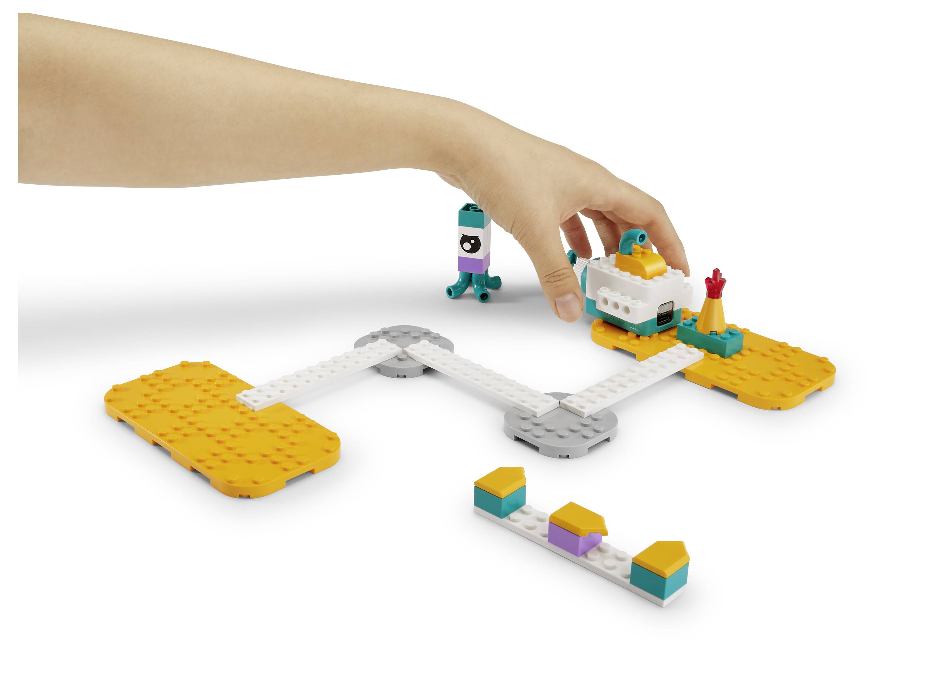 Kinderhand, die eine modulare Spielzeugstrecke mit kleinem Roboter und Blöcken auf weißem Hintergrund anordnet.