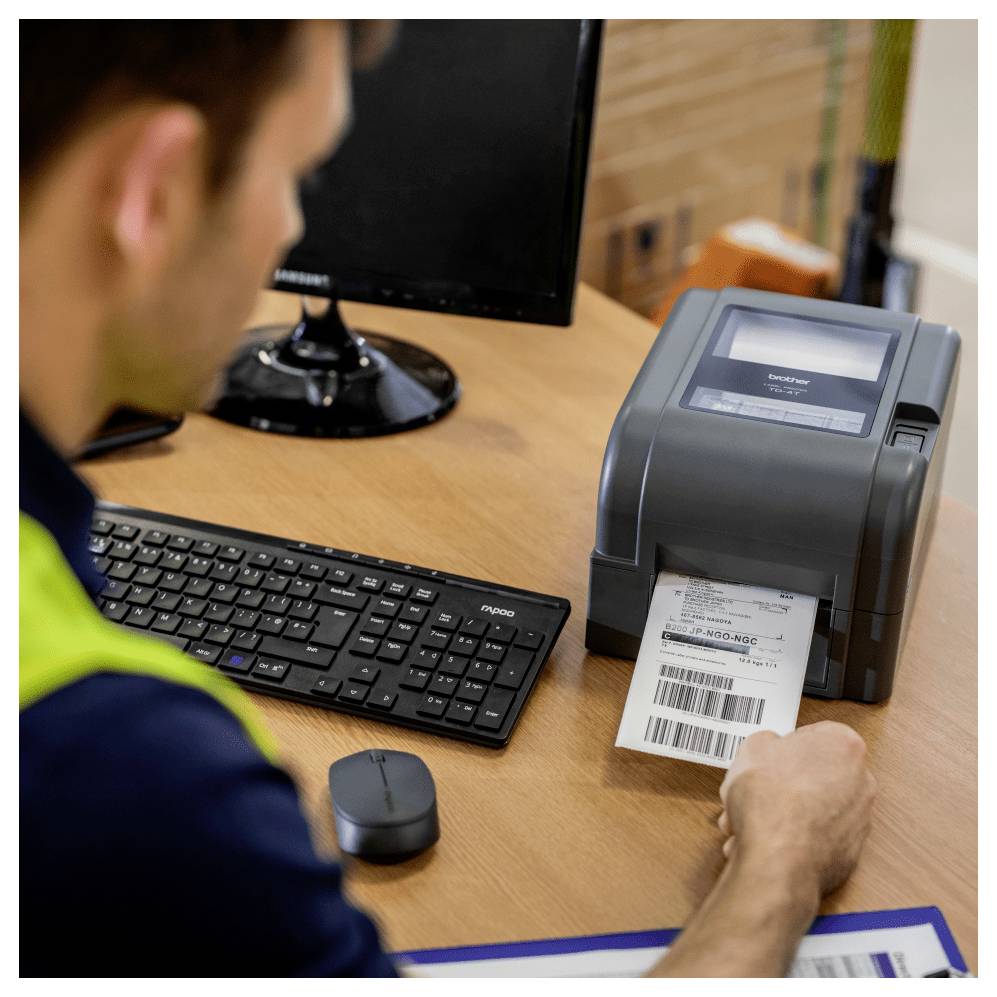 Arbeiter an einem Schreibtisch, der eine Barcode-Versandetiketten aus einem Etikettendrucker neben einem Computer und einer Tastatur druckt.