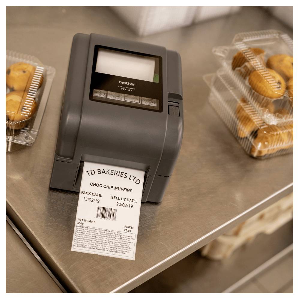 Etikettendrucker druckt ein Backwareenetikett für "TD Bakeries Ltd" Schokochip-Muffins auf einer Edelstahltheke, mit verpackten Muffins in der Nähe.