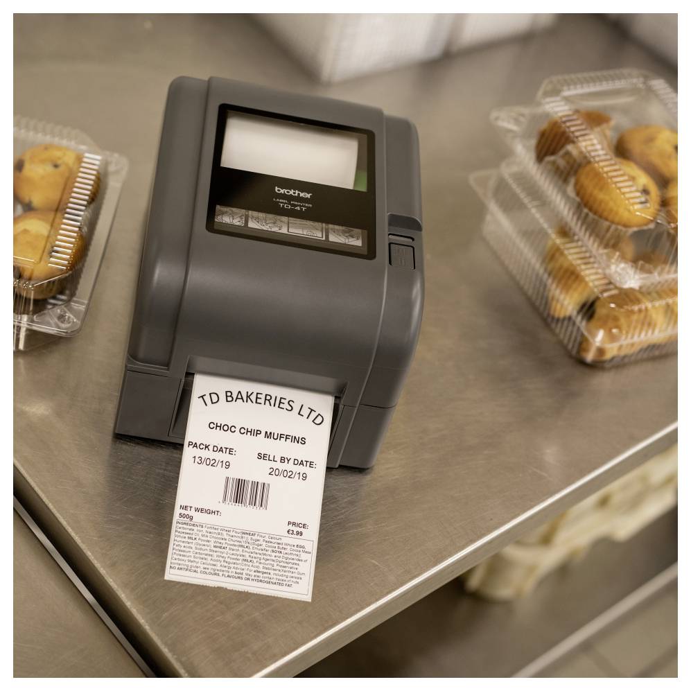 Etikettendrucker auf einer Bäckereitheke druckt ein Produktetikett für "TD BAKERIES LTD" Schokochip-Muffins, mit verpackten Muffins in der Nähe.