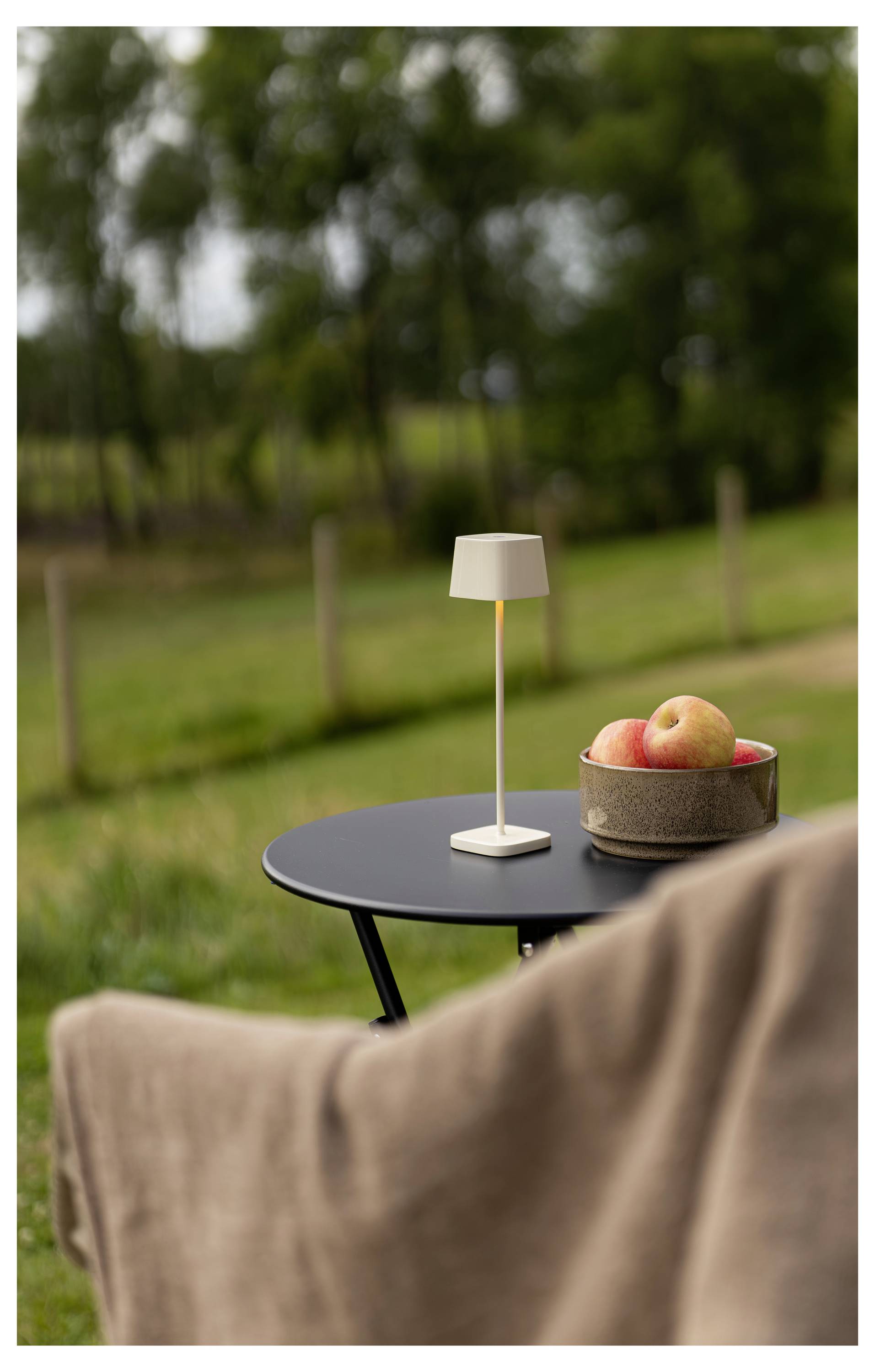 Kleiner Außentisch mit einer weißen Lampe und einer Schüssel mit Äpfeln neben einem unscharfen Stuhl in einer Gartenumgebung.