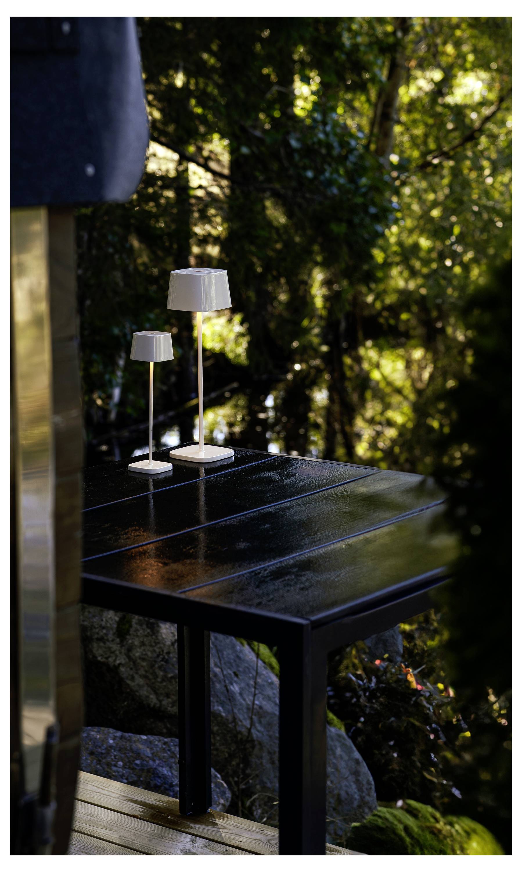 Zwei moderne Tischlampen, die auf einem schwarzen Außentisch auf einer Terrasse leuchten, mit unscharfem grünem Gartenhintergrund.