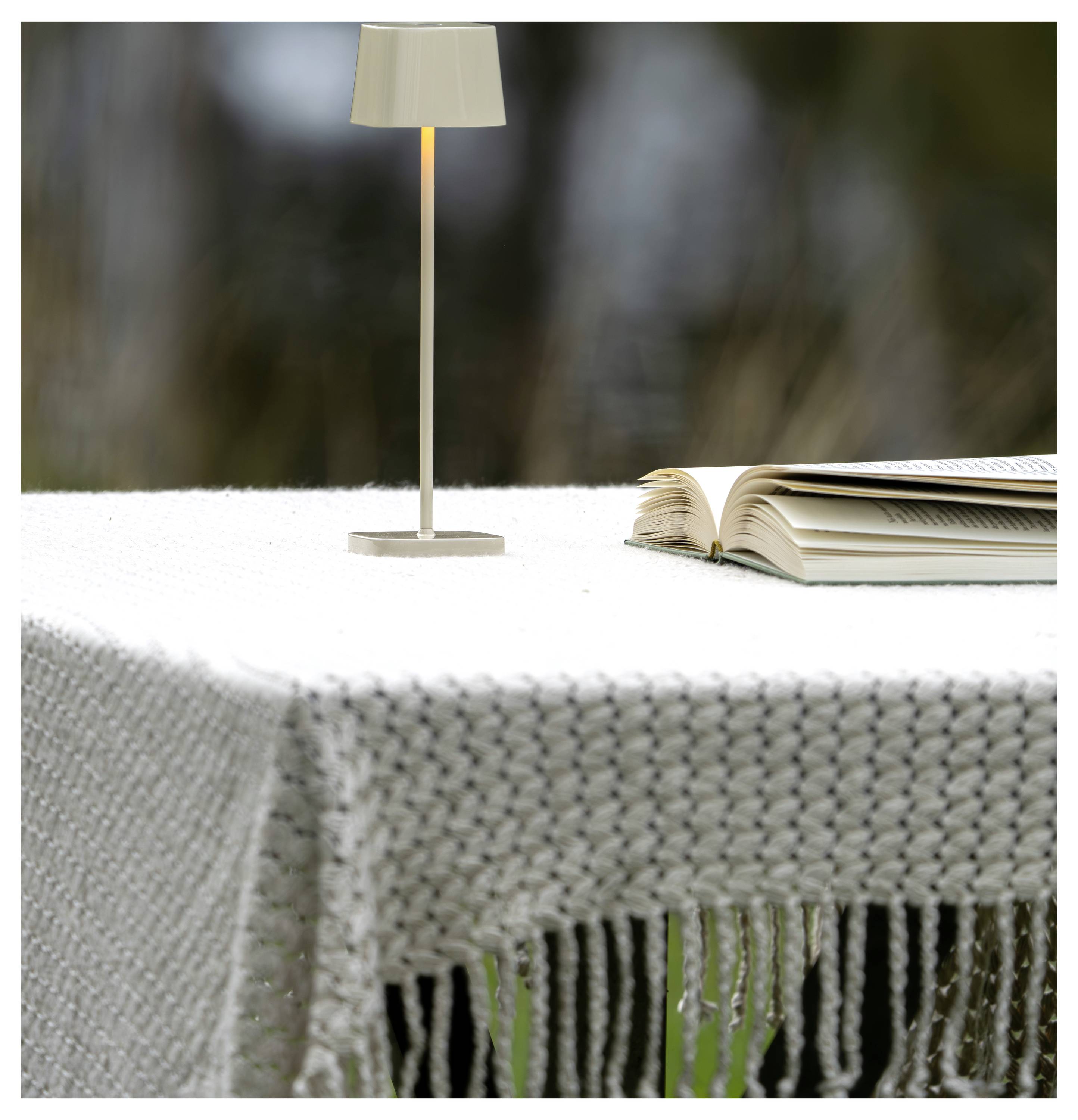 Ein offenes Buch neben einer kleinen Tischlampe sitzt auf einer weißen gewebten Tischdecke im Freien.