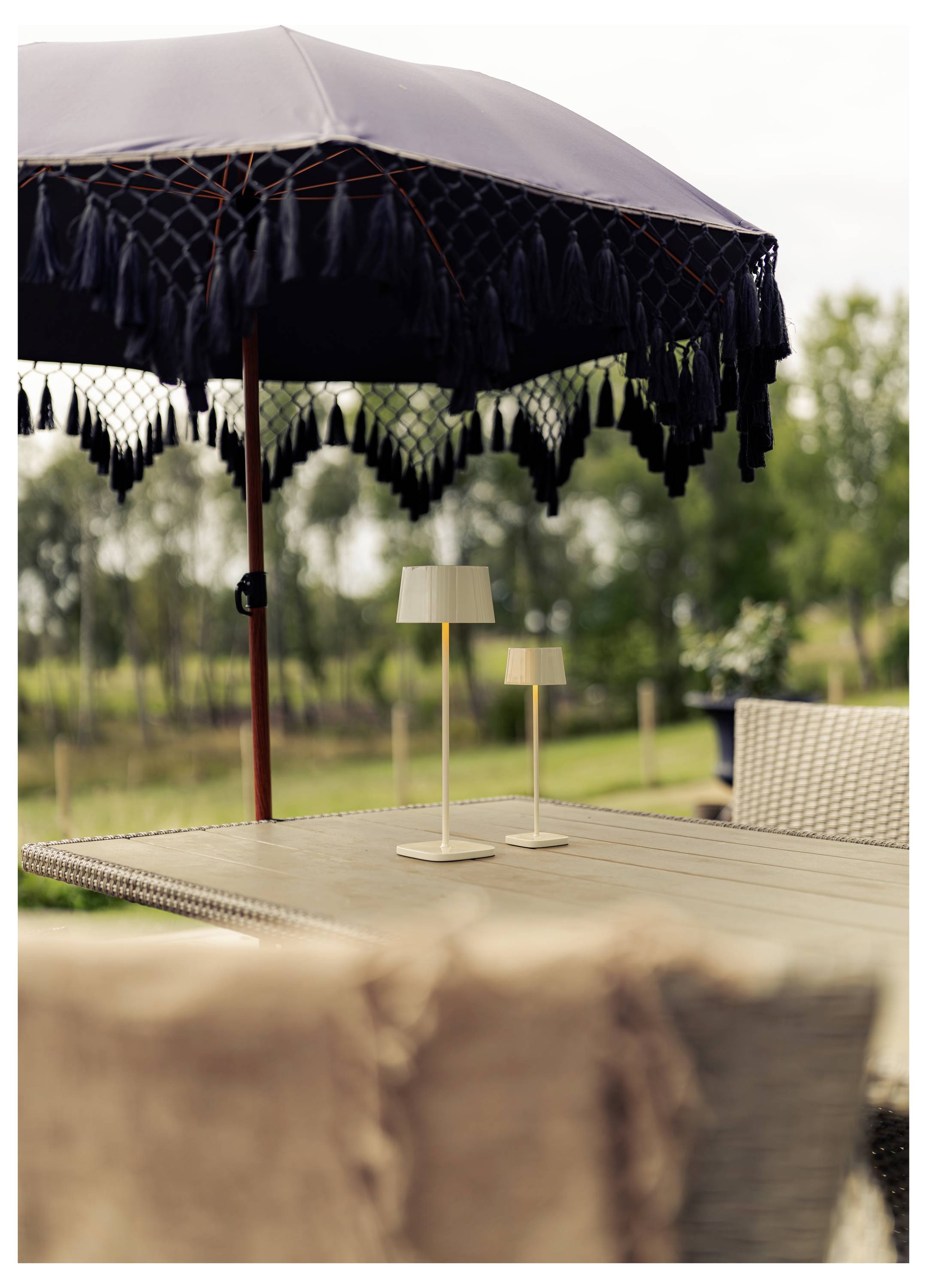 Sitzbereiche einer Außenterrasse mit einem großen Regenschirm mit Fransen und zwei kleinen Tischleuchten auf einem Holztisch mit Blick auf einen Garten.