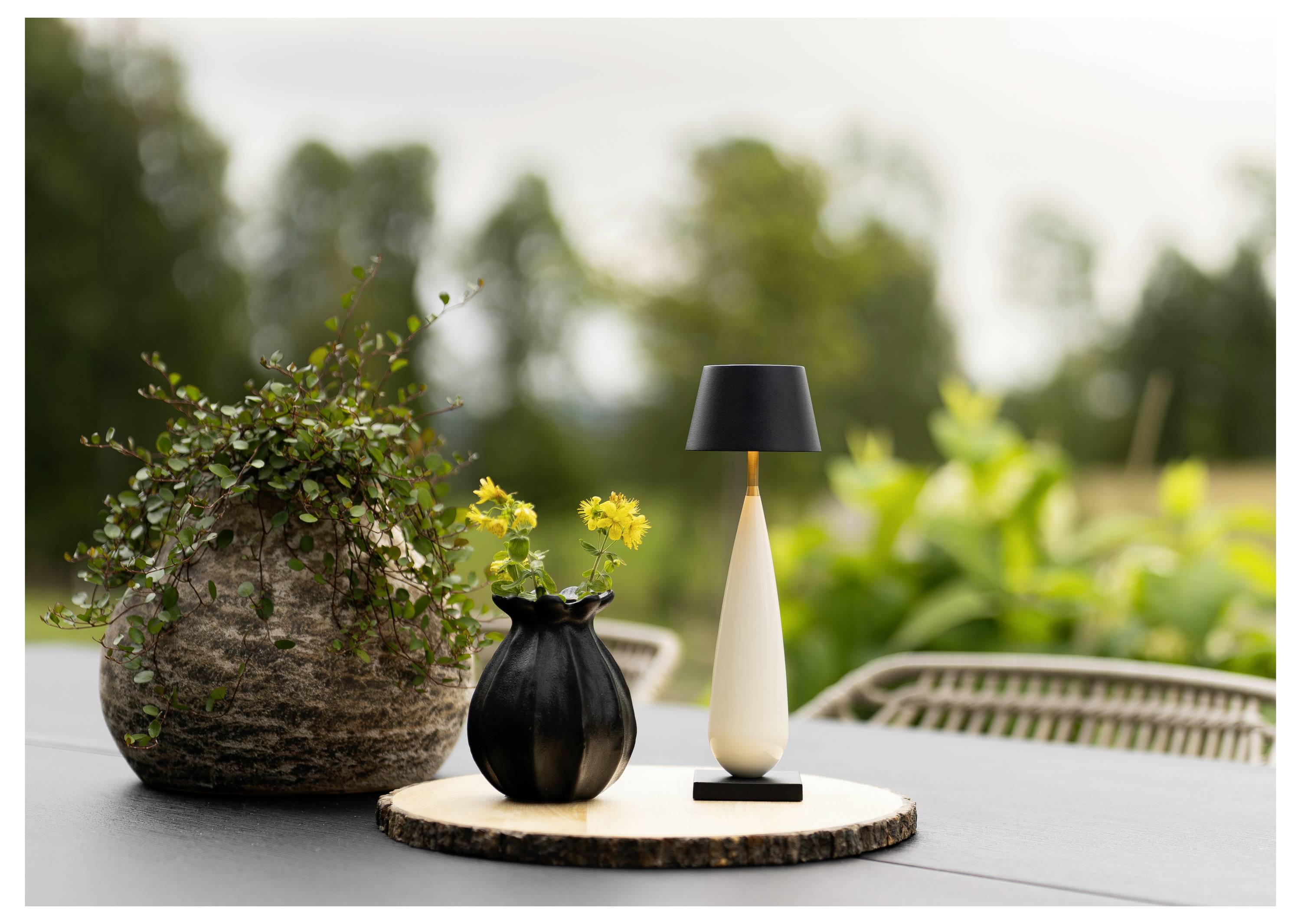 Outdoor-Tischdisplay mit einem großen Blumenkübel, einer kleinen schwarzen Vase mit gelben Blumen und einer modernen Lampe auf einem runden Holztablett mit Gartenhintergrund.