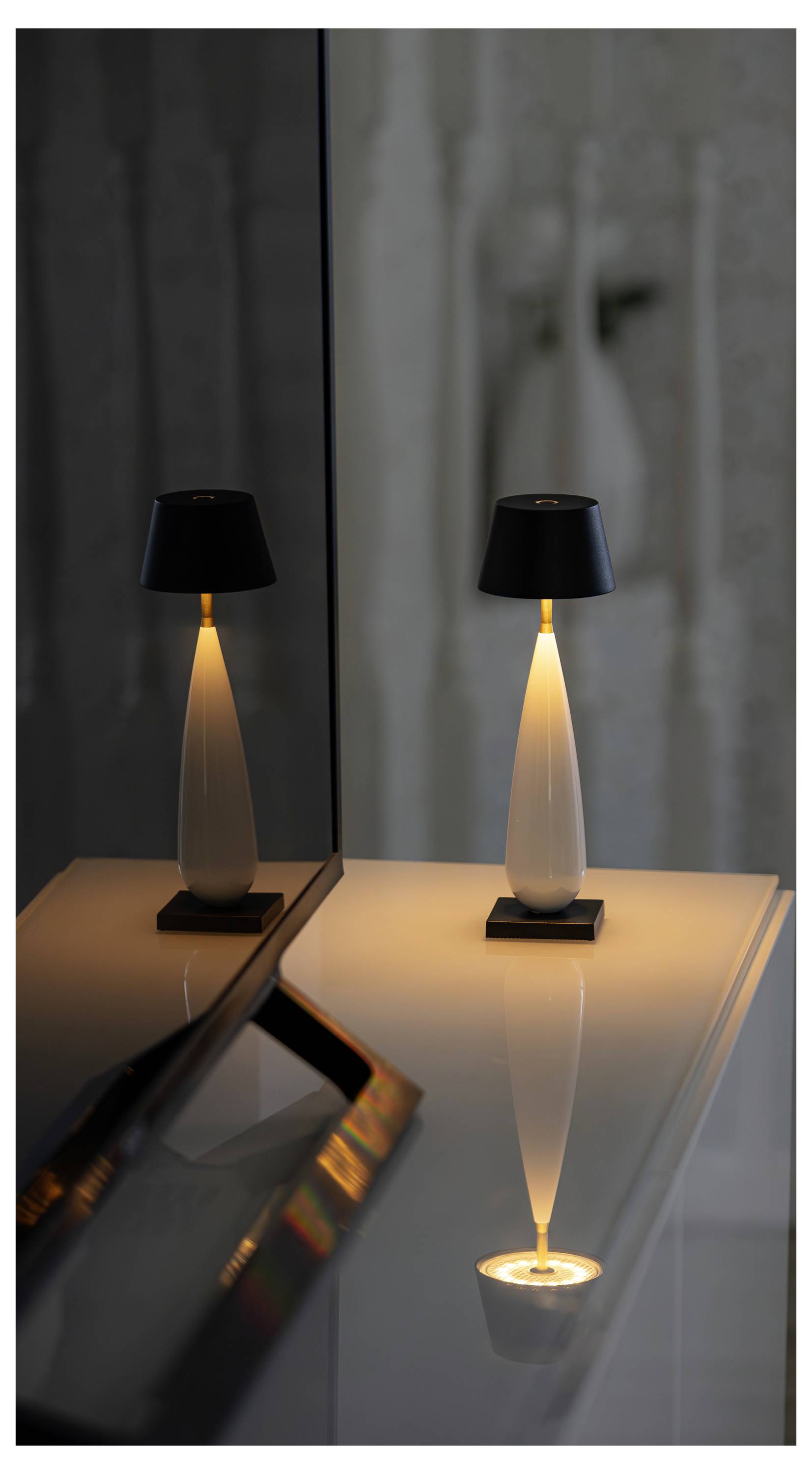 Tischlampe mit schwarzem Schirm und warmem Licht, das sich auf einer glänzenden Spiegelfläche in einem dunklen Raum widerspiegelt.
