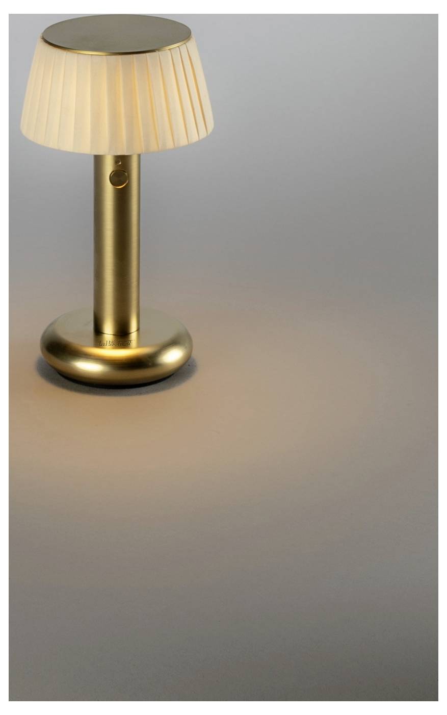 Eine kleine goldene Tischlampe mit einem gefalteten cremefarbenen Schirm wirft einen warmen, kreisförmigen Glanz auf eine graue Oberfläche.