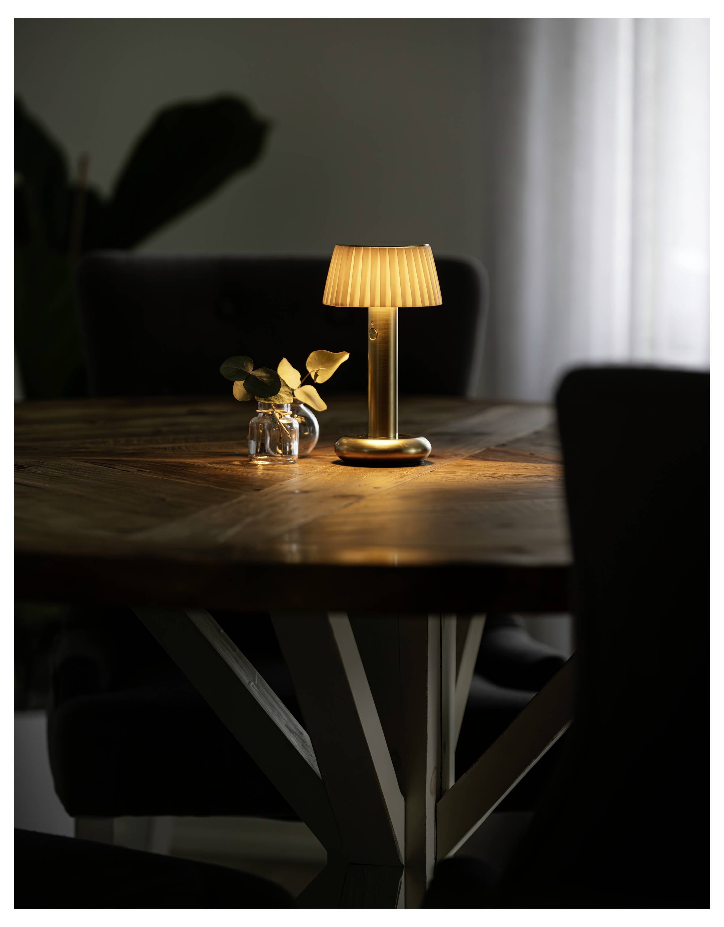 Eine warme Tischlampe leuchtet auf einem Holzesstisch neben einer kleinen Vase mit Blättern in einem dämmrigen Raum.