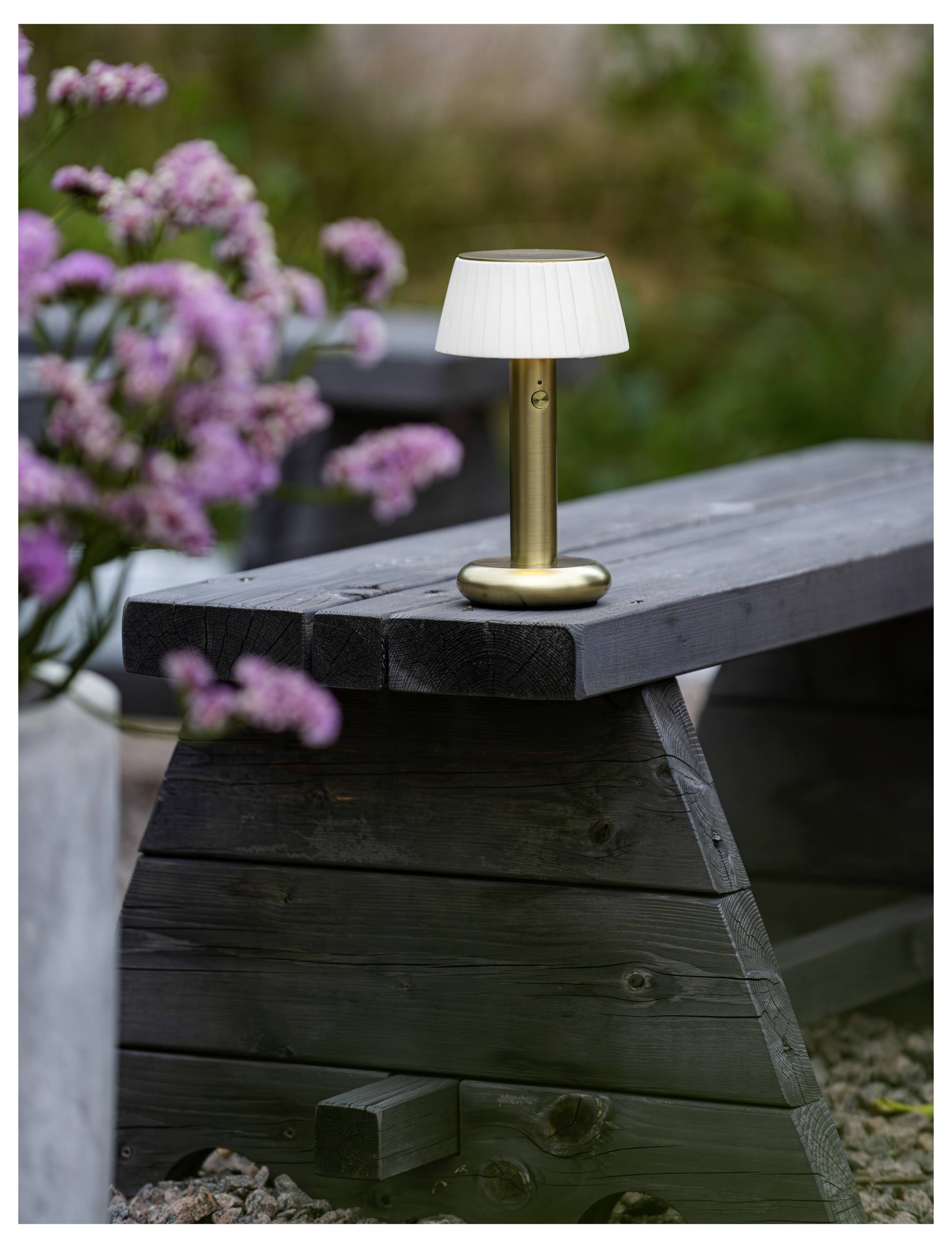Kleine Messingtischlampe mit weißem Schirm, die auf einer dunklen Holzbank im Freien steht, mit unscharfen rosa Blumen im Hintergrund.